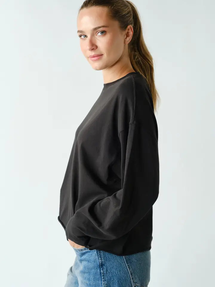 A woman wearing a black long-sleeve crewneck sweatshirt and blue jeans, shown from a side profile against a light gray studio background.