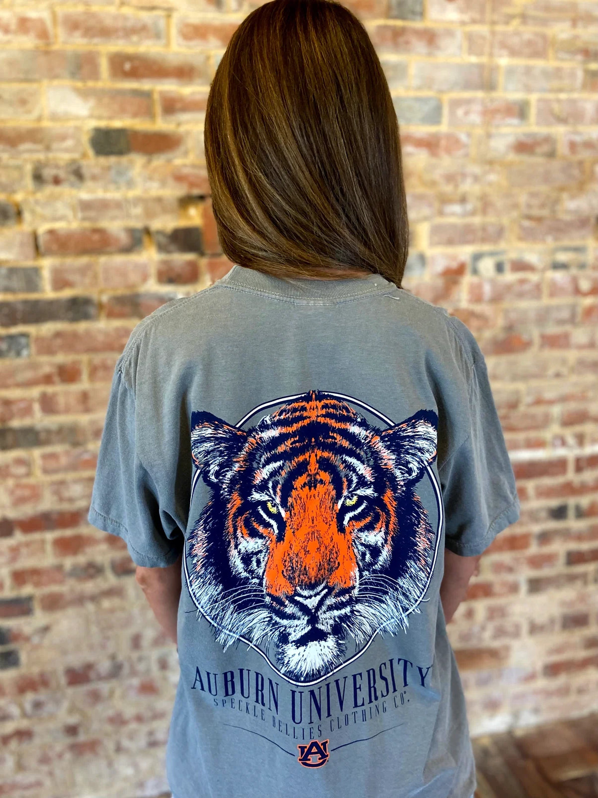 A woman viewed from behind wearing a faded gray t-shirt with a large orange and blue Auburn University tiger graphic on the back, standing in front of a brick wall.