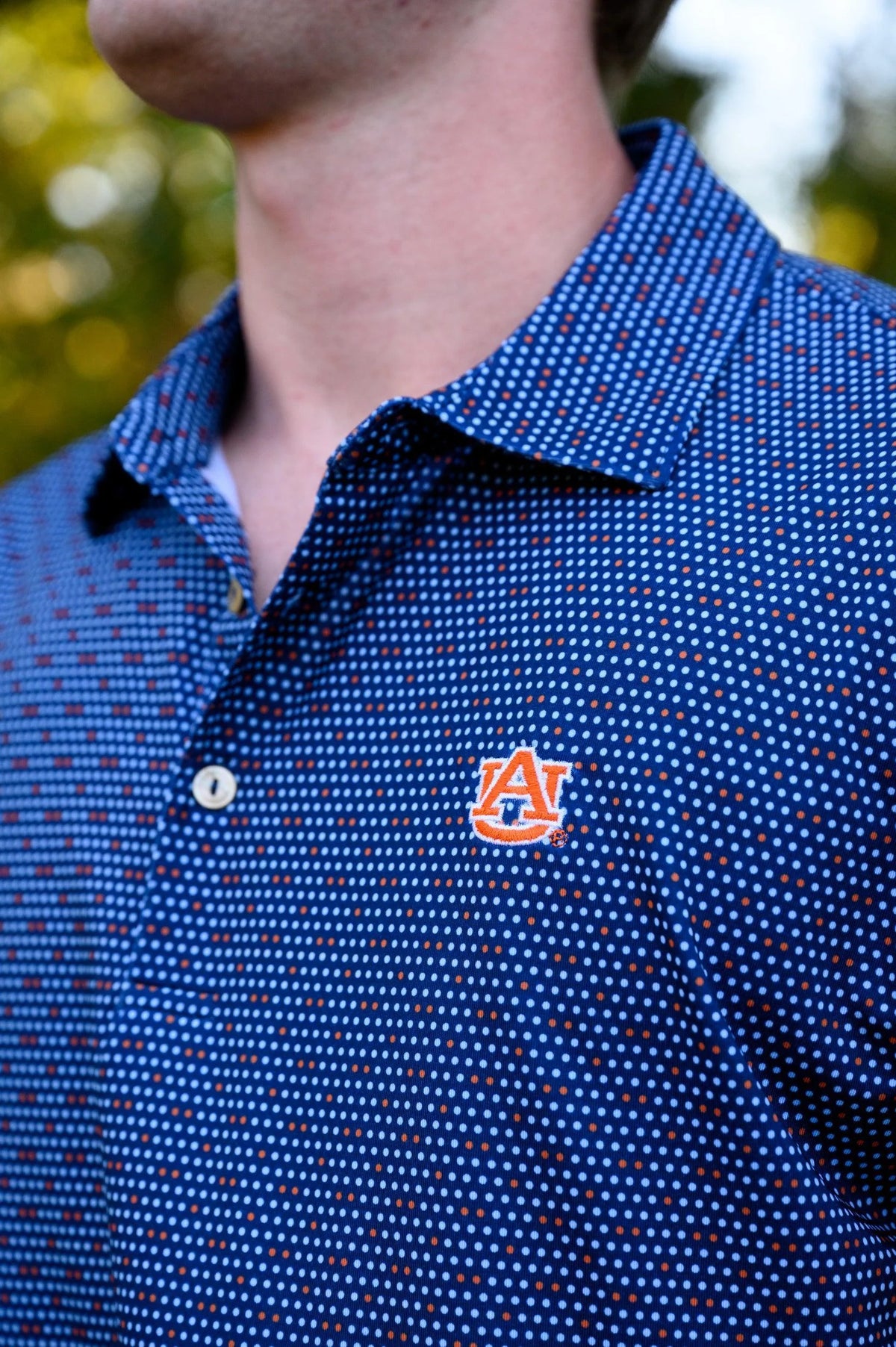 Close-up of a person wearing a blue short-sleeve polo shirt with a dense small white and red dot pattern and an Auburn Tigers 'AU' logo patch on the left chest, photographed outdoors with blurred green foliage in the background.