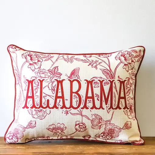 A rectangular decorative throw pillow with a cream fabric base and red floral pattern, featuring the word 'ALABAMA' in large red letters across the front, resting on a wooden surface against a white wall.