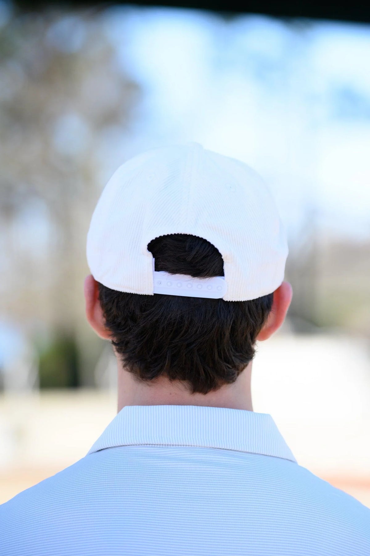 A man viewed from behind wearing a white baseball cap with the strap undone, a light blue collared shirt, and dark brown hair. The background is a soft-focus outdoor scene.