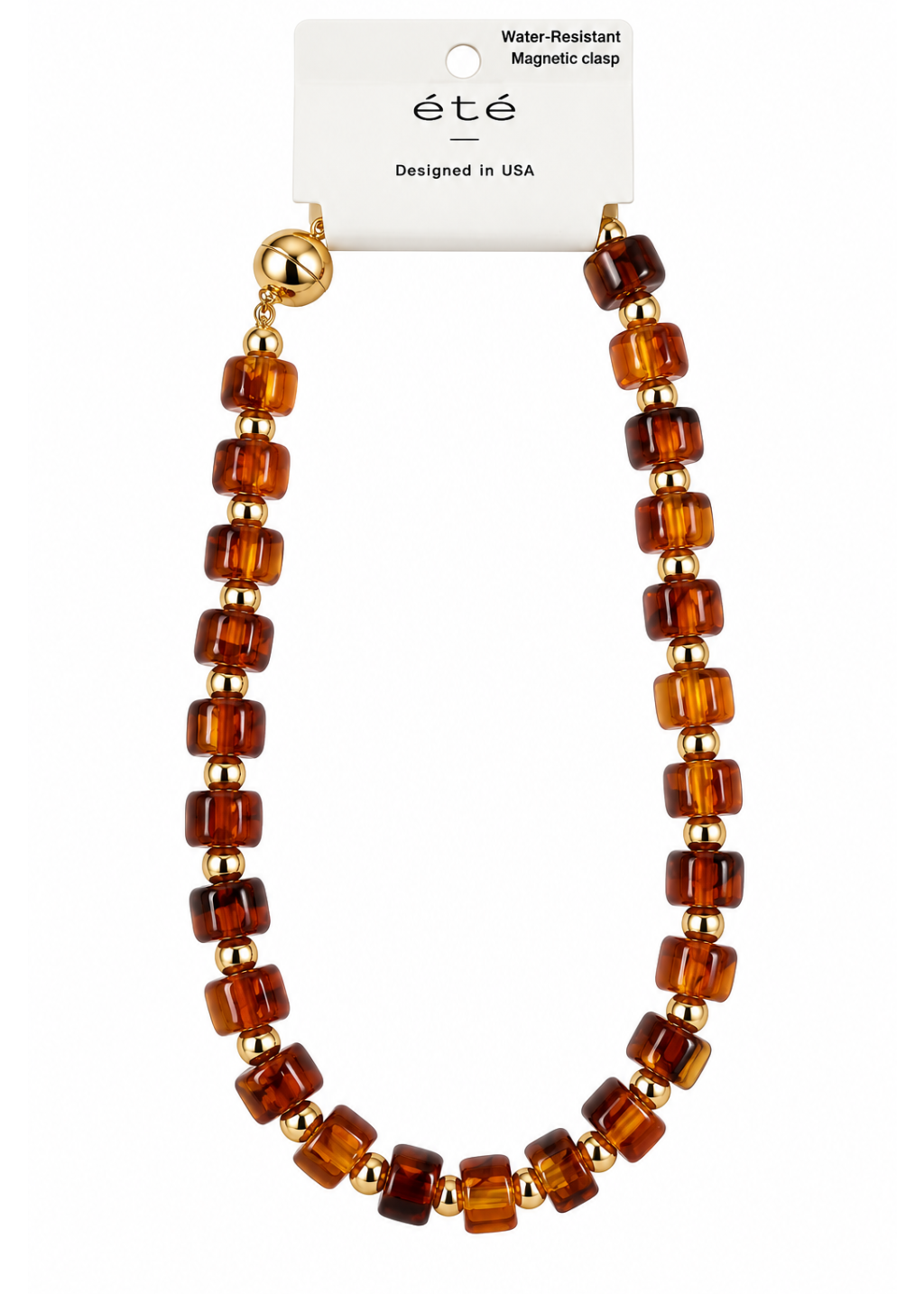 A water-resistant magnetic clasp necklace made of amber-brown translucent beads separated by small gold spacer beads, hanging from a white card labeled “été” with text reading “Water-Resistant Magnetic clasp” and “Designed in USA.”.