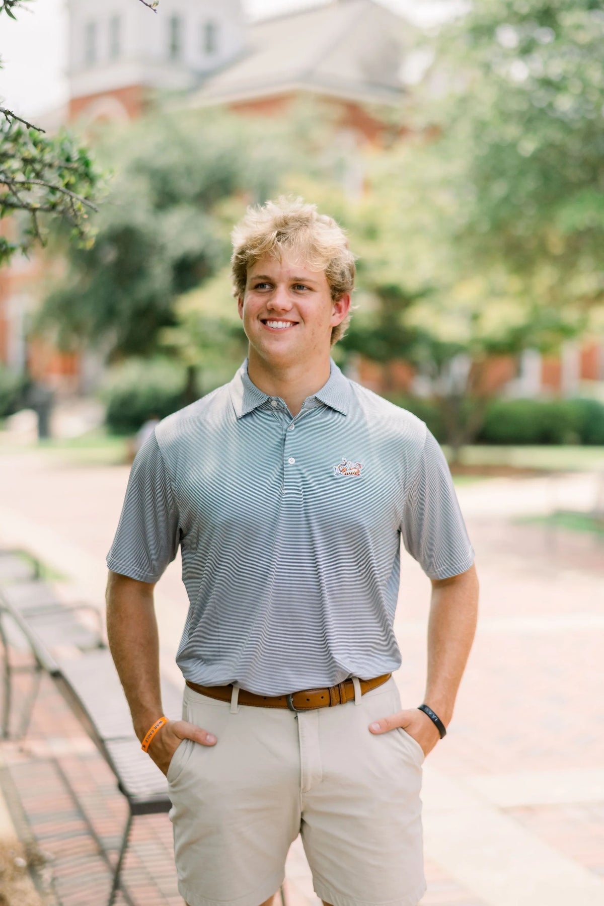 A smiling young man standing outdoors on a campus walkway, wearing a light blue polo shirt with a small embroidered logo on the left chest, khaki shorts, and a brown belt.