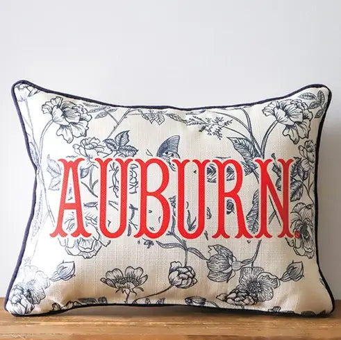 A rectangular decorative throw pillow with a navy blue floral vine pattern on a cream background, featuring the word 'AUBURN' in large red letters across the front, resting on a wooden surface against a light gray wall.