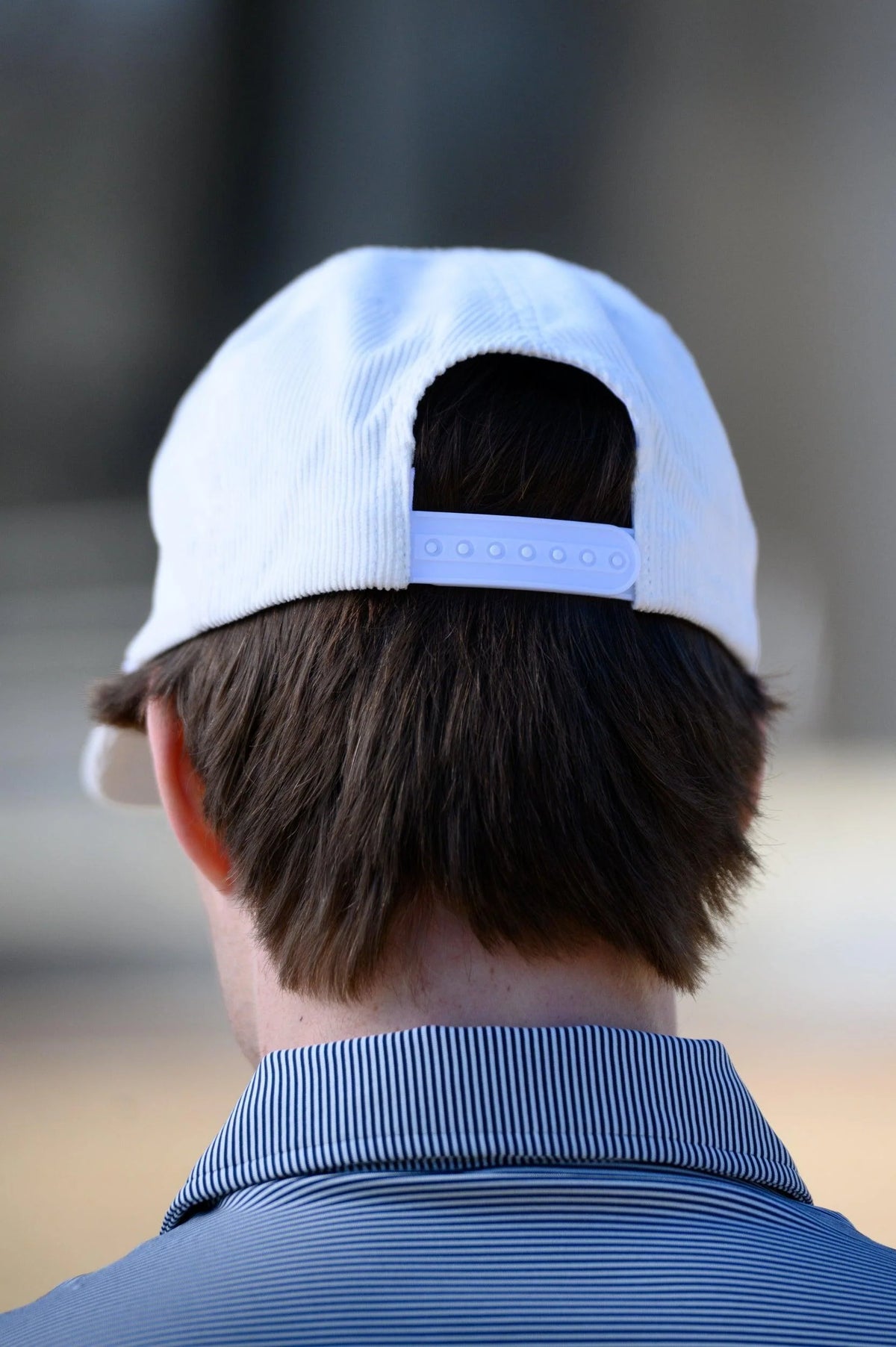 Back view of a man wearing a white corduroy baseball cap with an adjustable strap and a blue striped polo shirt.