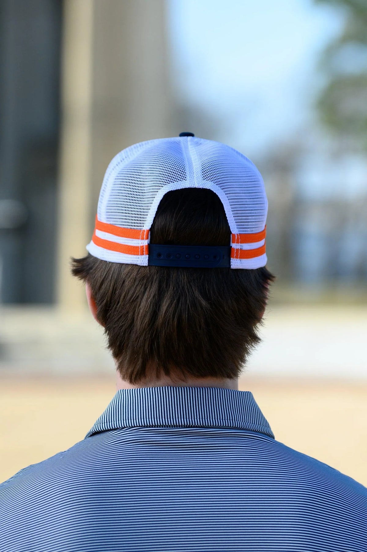 A man viewed from behind wearing a white mesh back trucker cap with orange stripes and a navy adjustable strap, paired with a blue striped polo shirt in an outdoor setting.