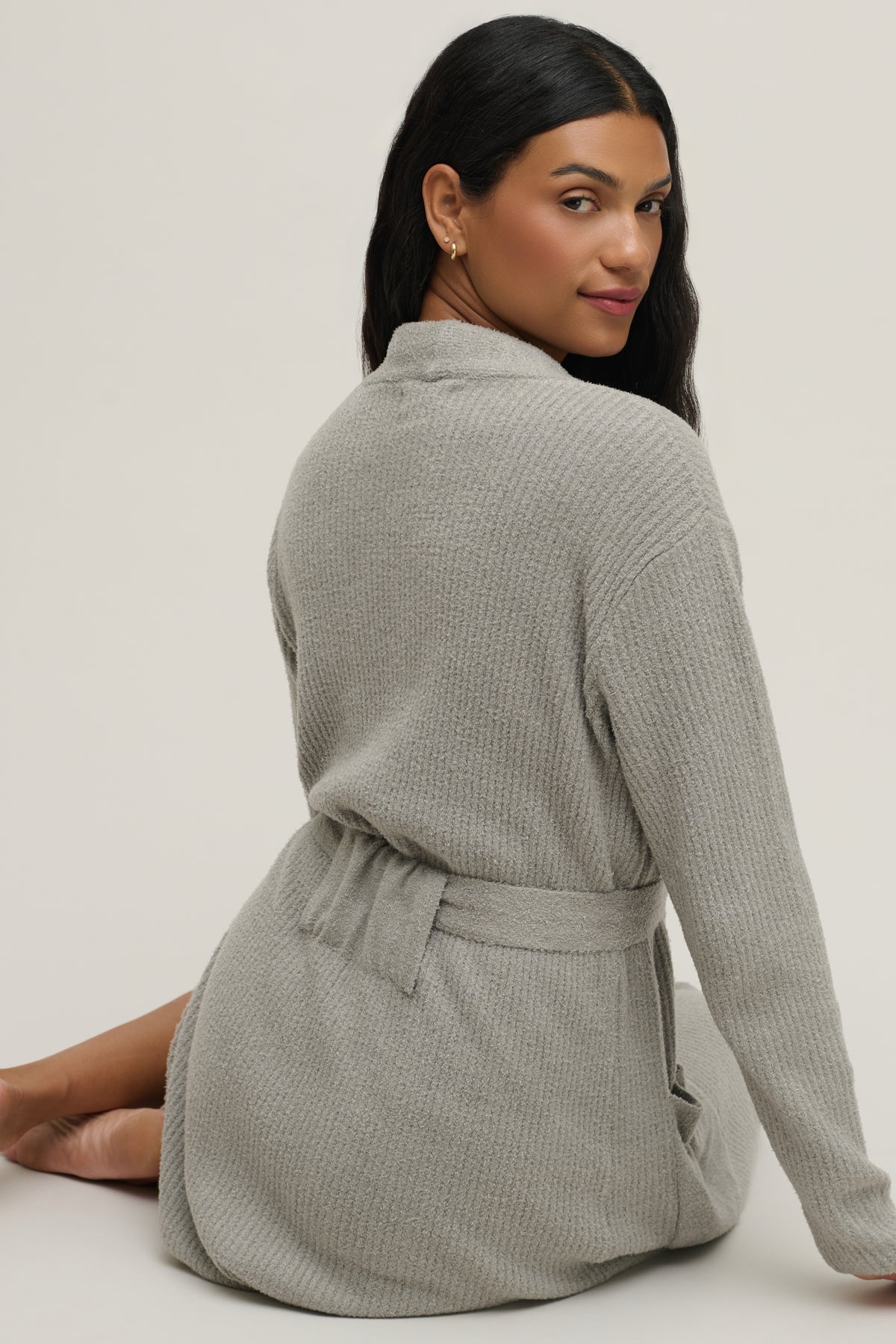 A woman wearing a light gray waffle-knit robe, shown from the back and side, sitting against a plain light gray studio background.