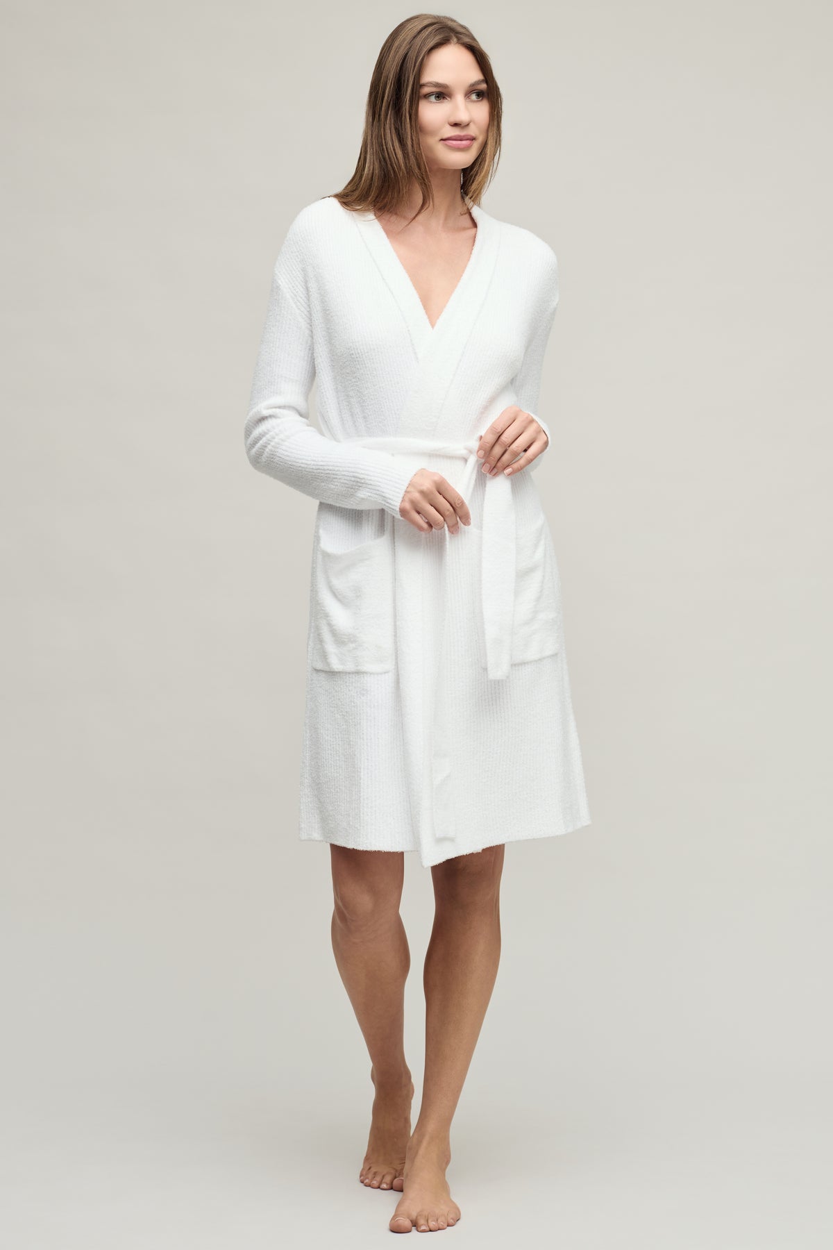 A woman modeling a white long-sleeve bathrobe with a V-neck, front pockets, and a self-tie belt, photographed against a plain light gray studio background.