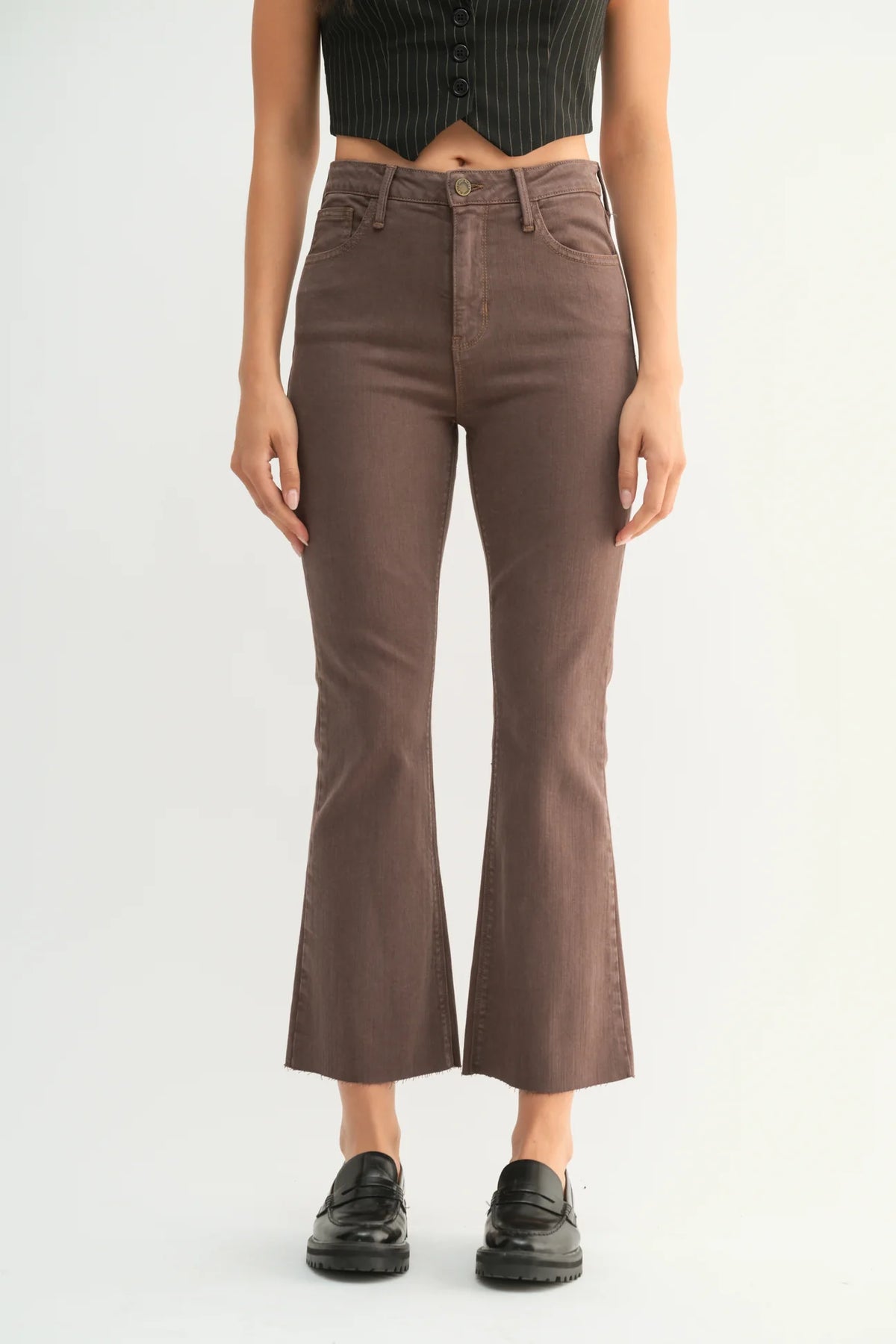 A model is shown from the waist down wearing brown cropped flare jeans, a black ribbed crop top, and black leather loafers against a plain white studio background.