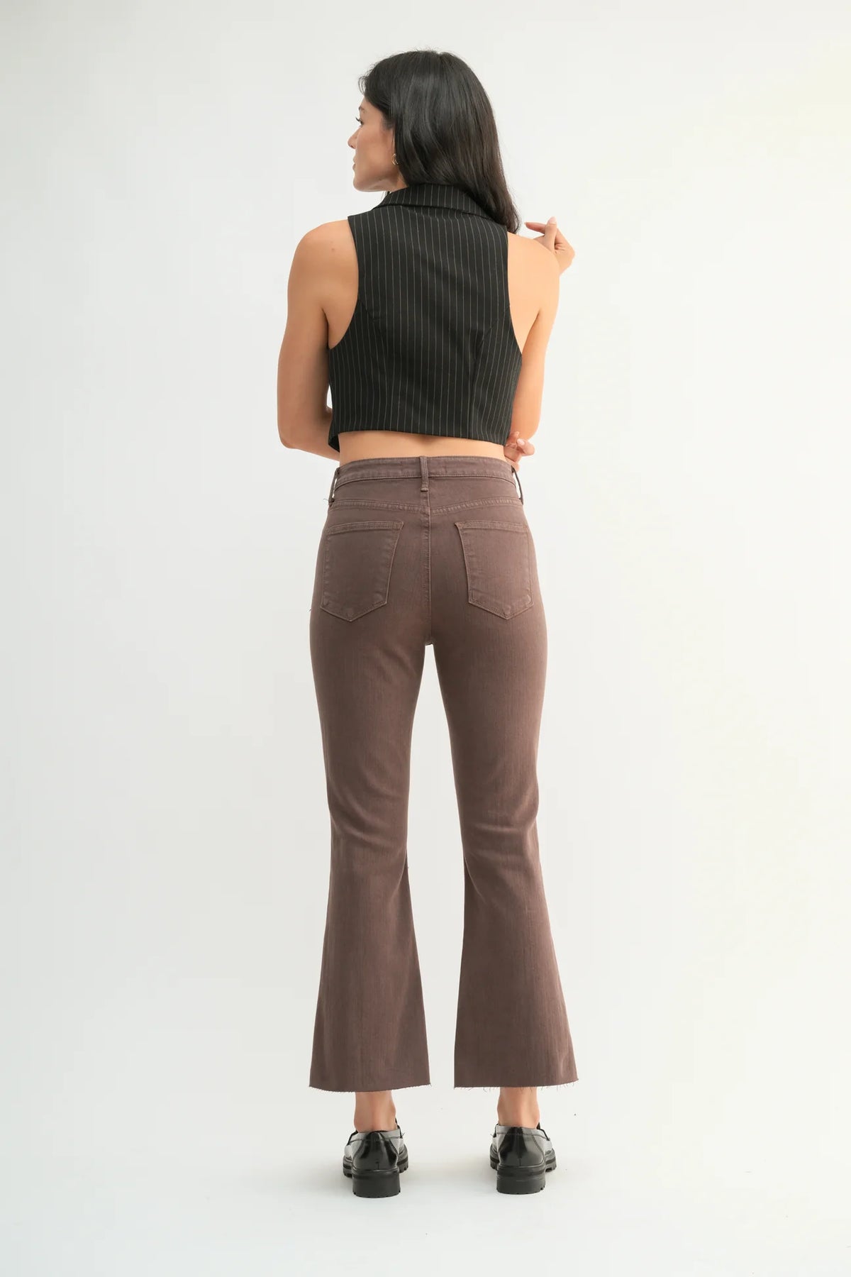A woman is shown from the back wearing brown flared pants, a black ribbed sleeveless crop top, and black shoes against a plain white studio background.