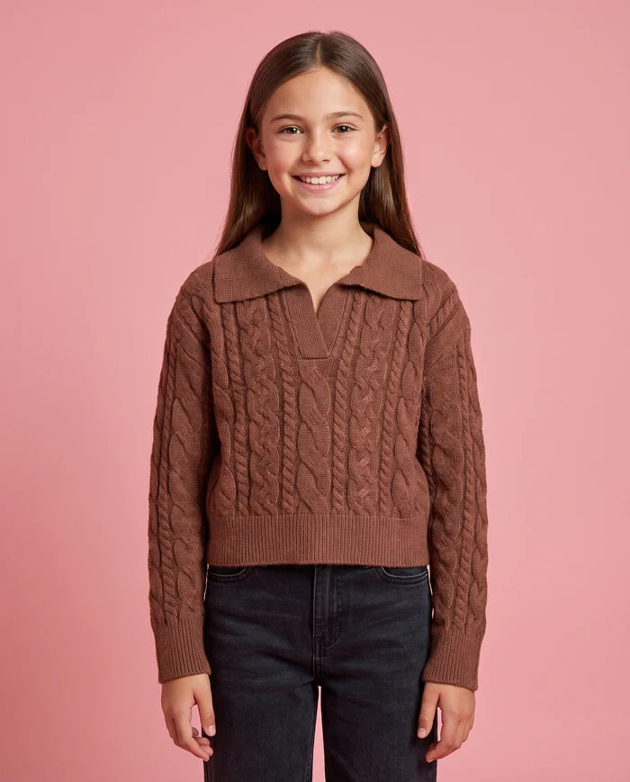Girl standing on pink background wearing a brown cable knit sweater and black jeans 