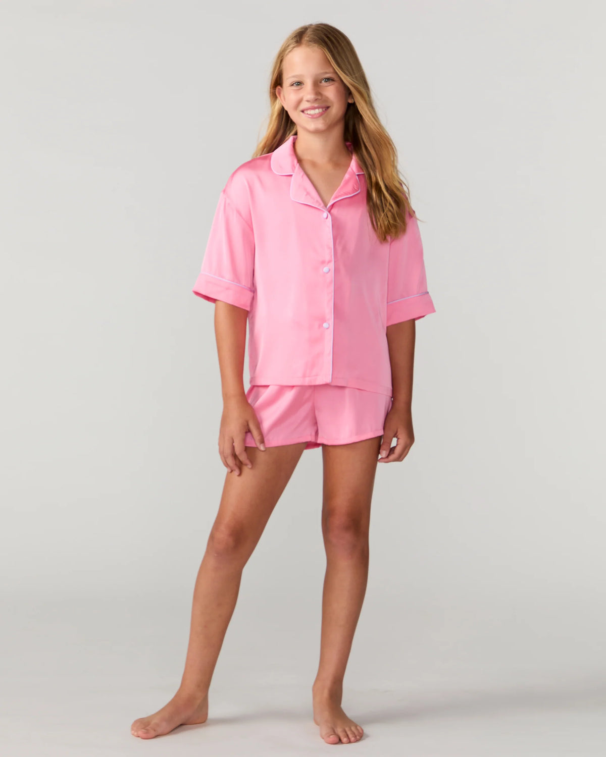 A young girl modeling a two-piece pink pajama set consisting of a short-sleeve button-up shirt and matching shorts, photographed against a neutral light gray studio background.