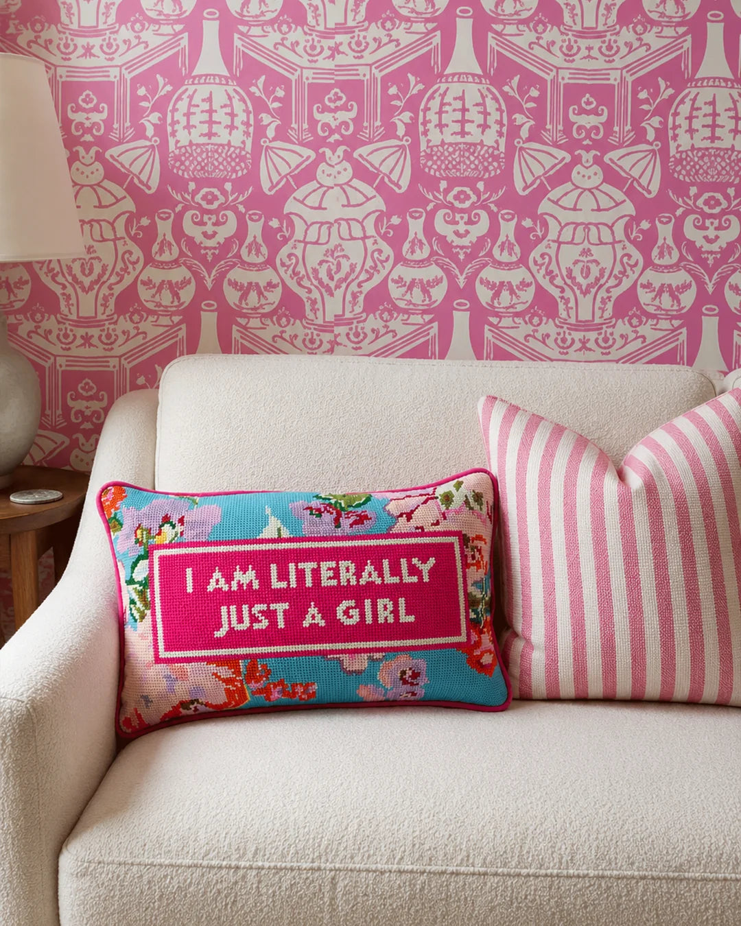 a floral needlepoint pillow with the words "i am literally just a girl" on it on a cream couch. decorated with pink wallpaper and pillows around