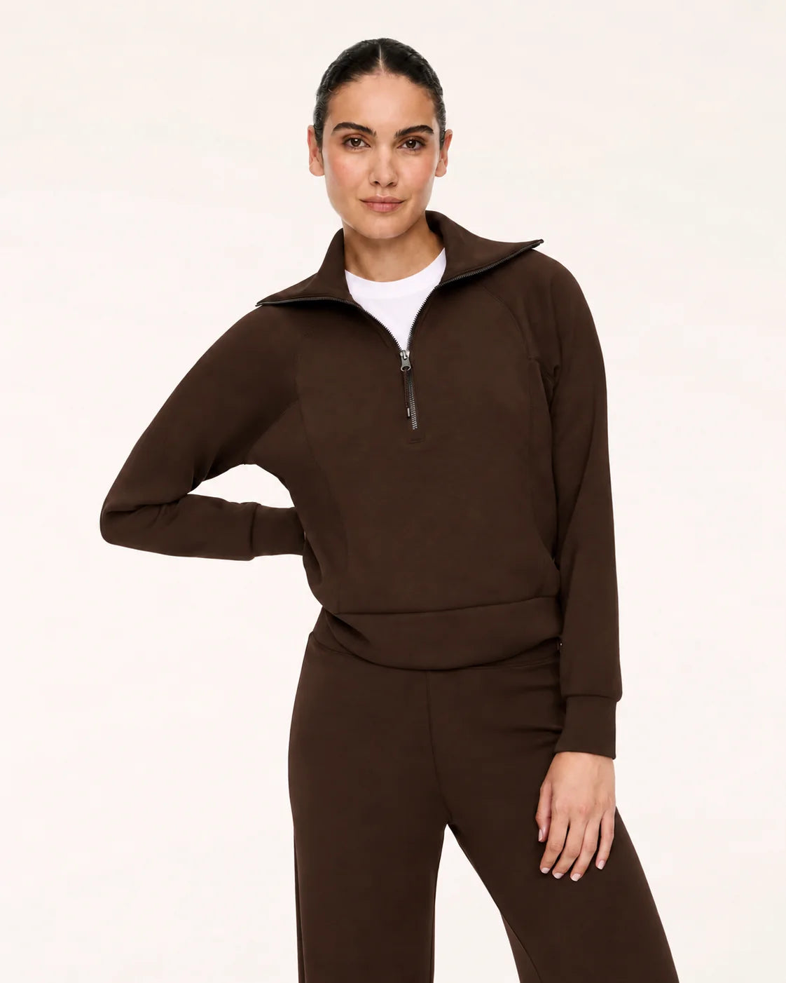 A woman modeling a dark brown long-sleeve quarter-zip pullover and matching dark brown pants over a white crew-neck undershirt, photographed against a clean off-white studio background.