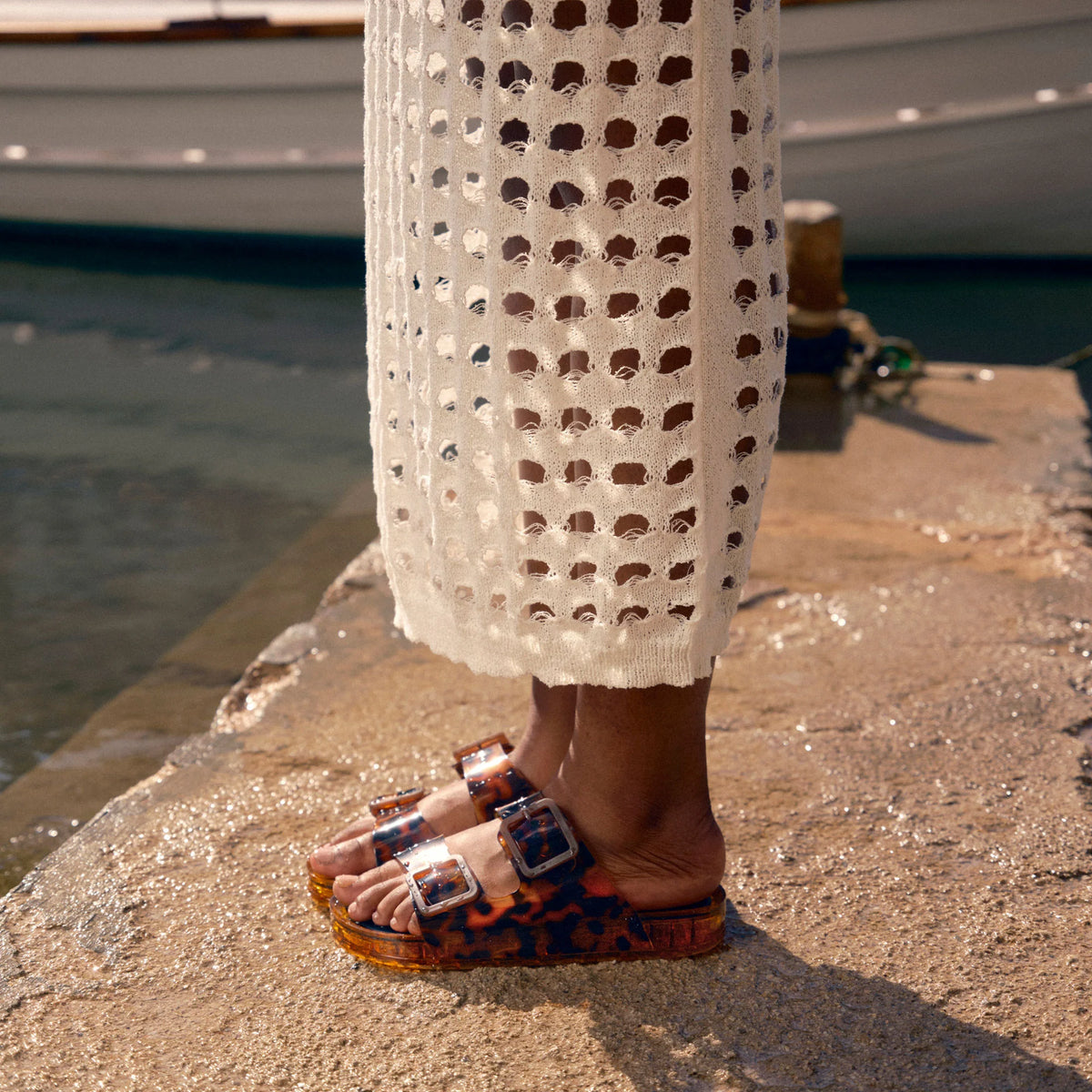 Person wearing a white crochet cover-up and patterned sandals by a waterfront.