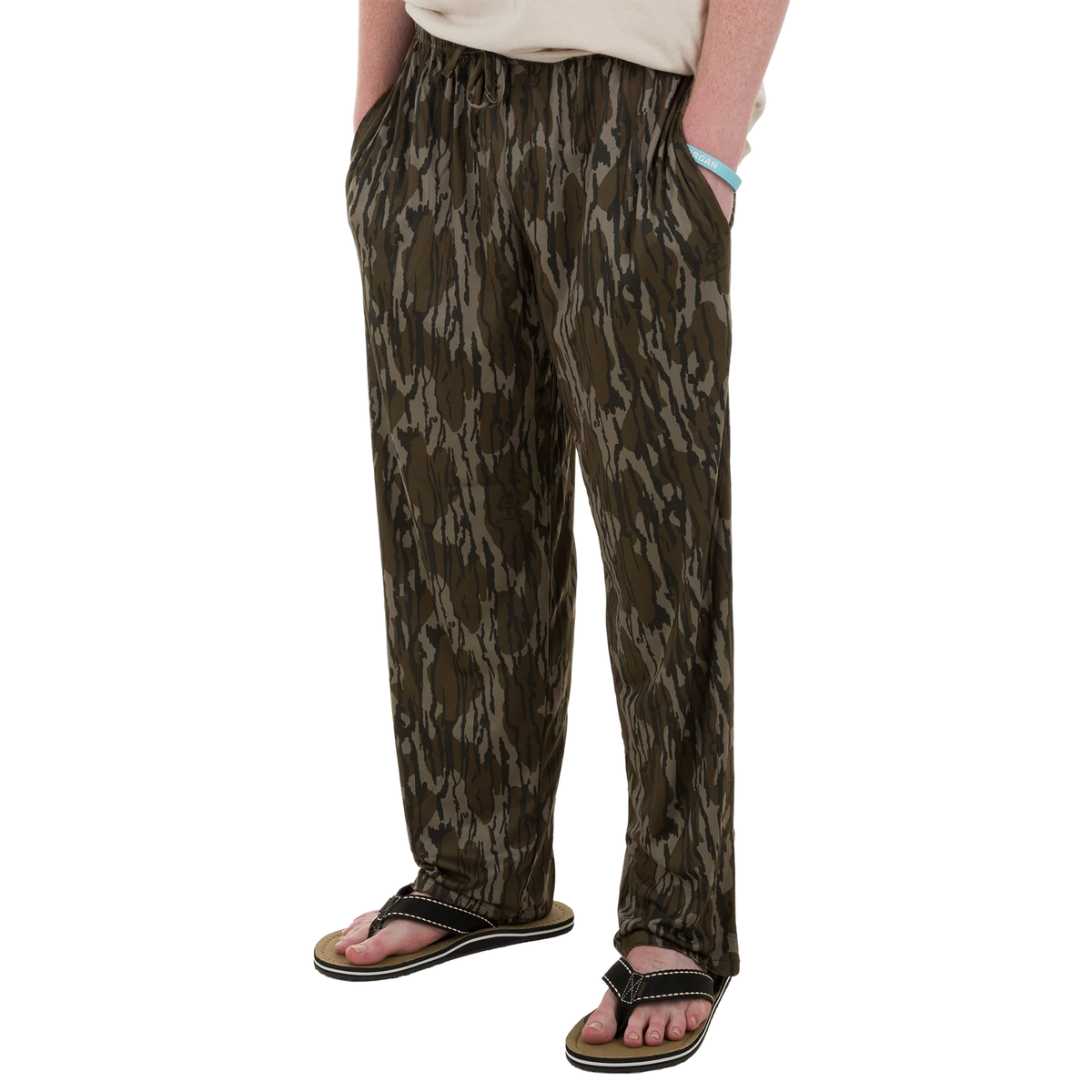 A person wearing loose, drawstring pants with an allover camouflage-like pattern in olive green, brown, and black, paired with black slide sandals against a plain white background.