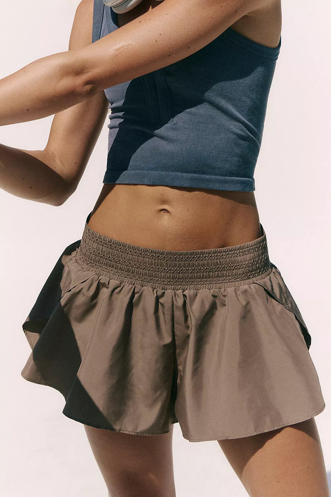 A close-up of a model wearing a dark slate blue cropped tank top and a pleated athletic skirt in taupe brown with dark side panels, set against a plain off-white background.