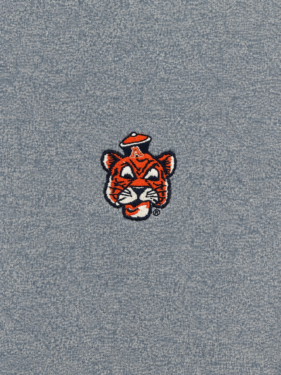 A close-up of a heather gray fabric surface featuring a small embroidered tiger mascot patch with orange, black, white, and red details.