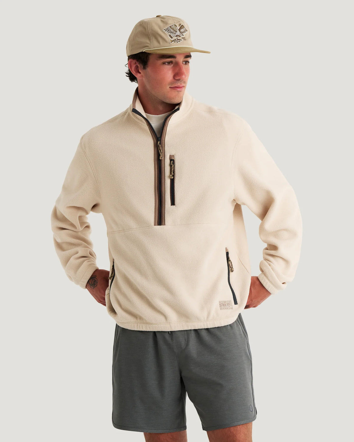 A man wearing a beige quarter-zip fleece pullover, gray shorts, and a beige baseball cap, standing against a light gray studio background.