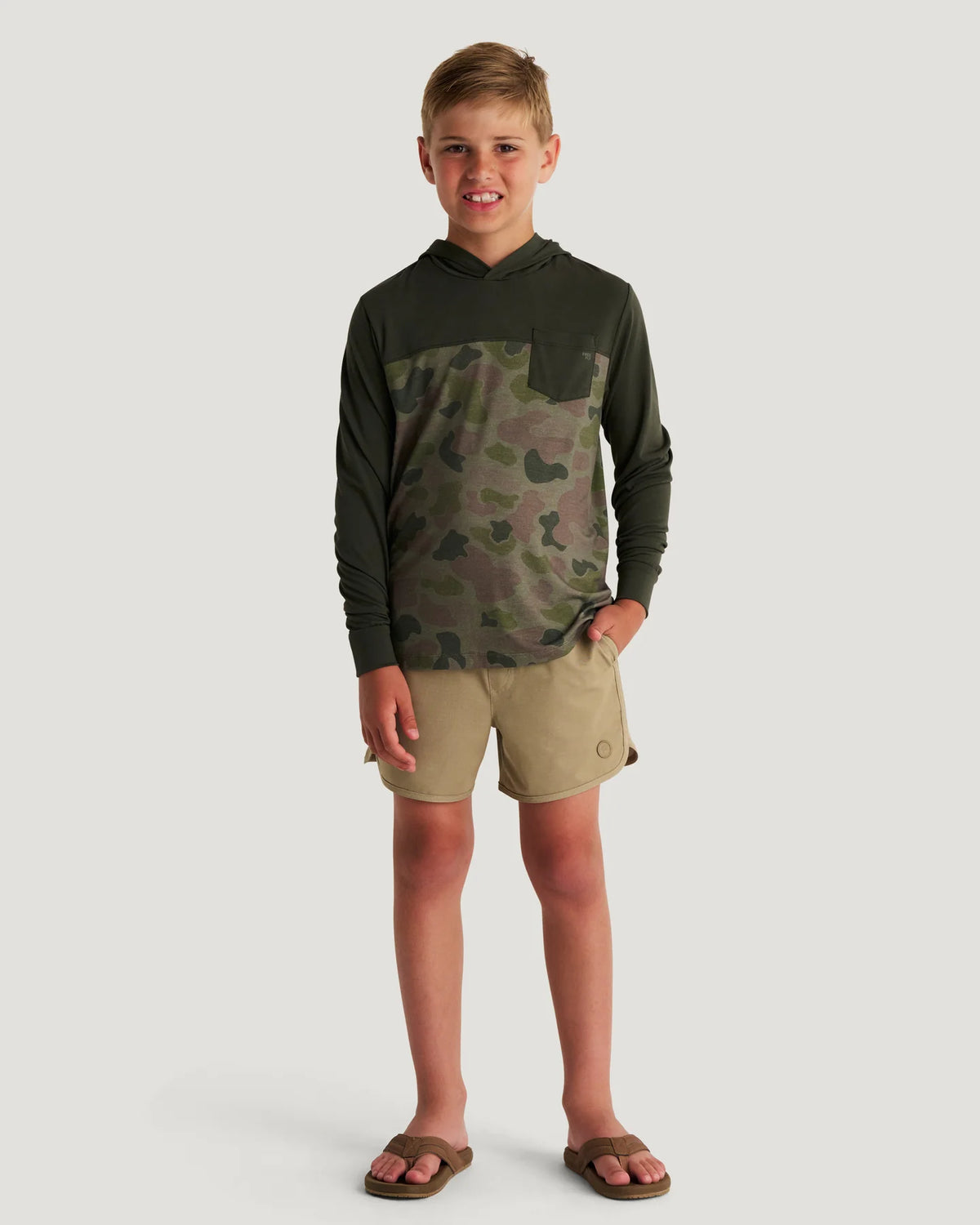 Boy wearing a green and camouflage hoodie and tan shorts with flip flops standing in front of a white background. 