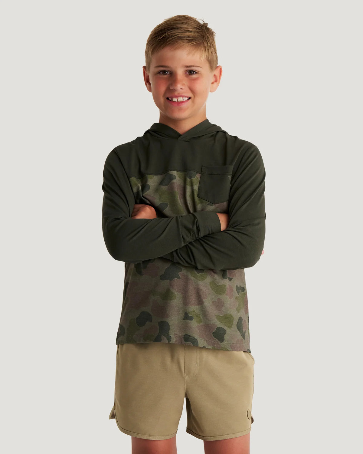 Boy wearing a green and camouflage hoodie and tan shorts with flip flops standing in front of a white background. 