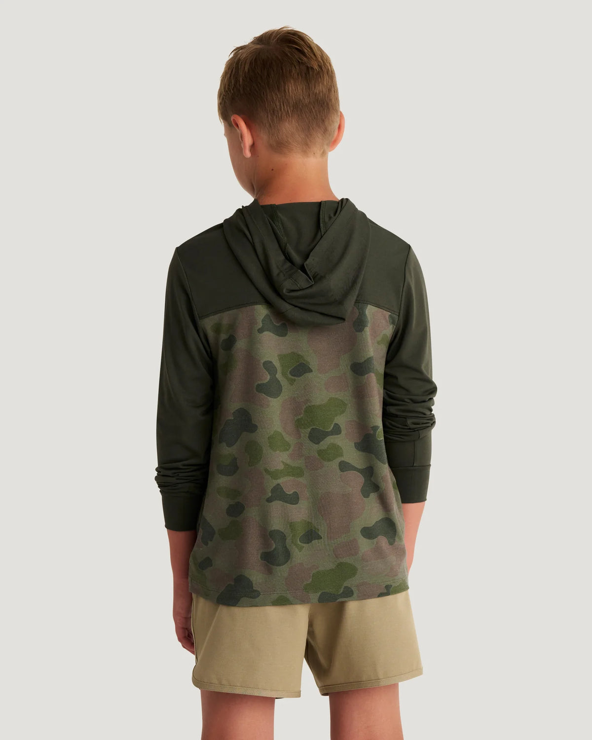 Boy wearing a green and camouflage hoodie and tan shorts with flip flops standing in front of a white background. 