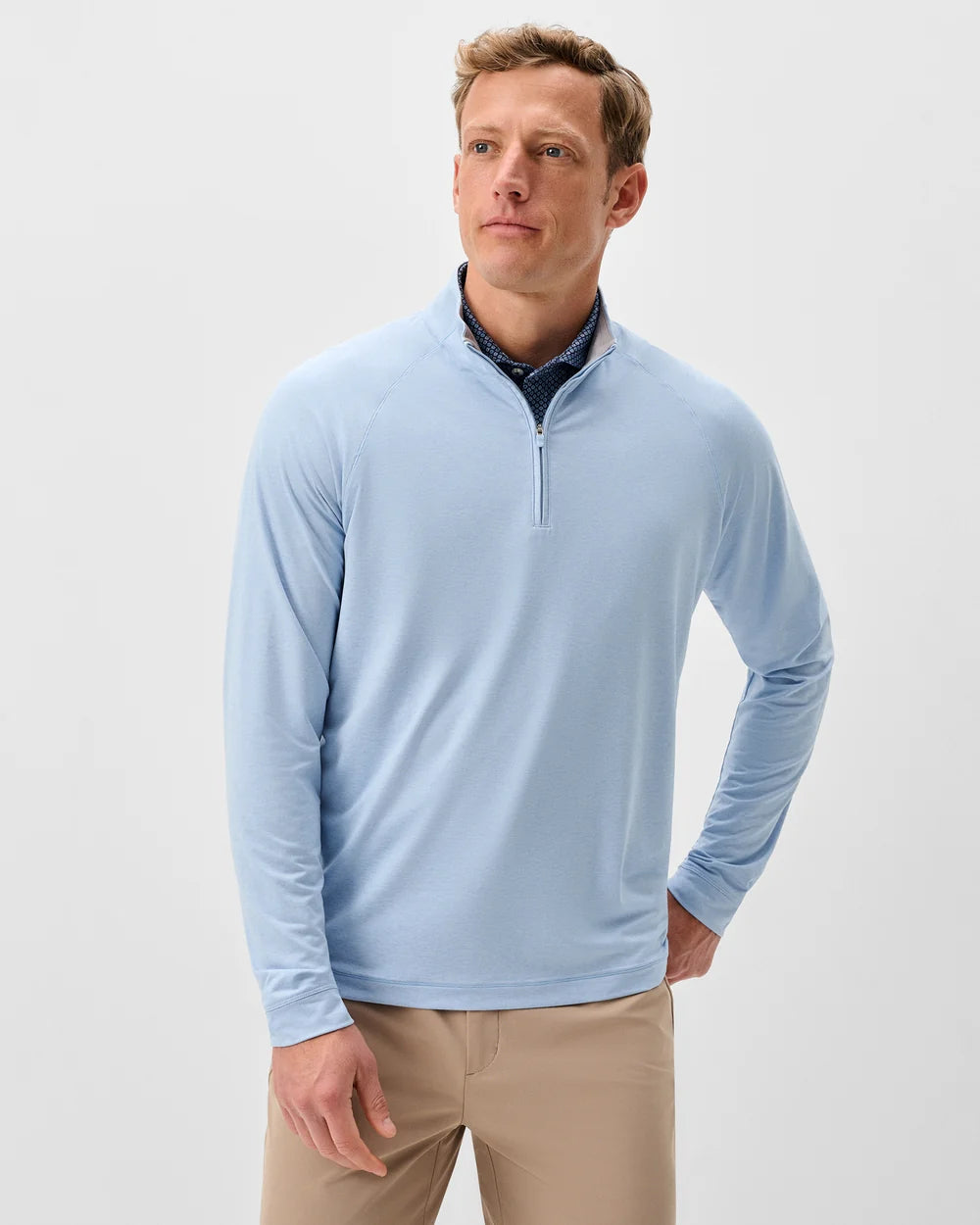 A man wearing a light blue long-sleeve quarter-zip pullover over a dark patterned collared shirt and khaki pants, photographed against a clean light gray studio background.