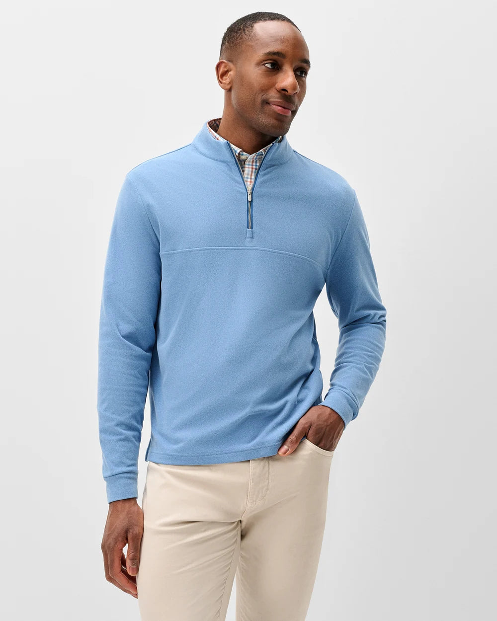 A man wearing a light blue long-sleeve quarter-zip pullover sweater over a patterned collared shirt and beige trousers, standing against a light gray studio background.