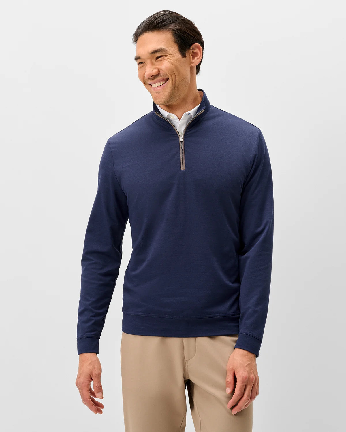 A smiling man wearing a navy long-sleeve quarter-zip pullover and khaki pants, standing in front of a white background. 