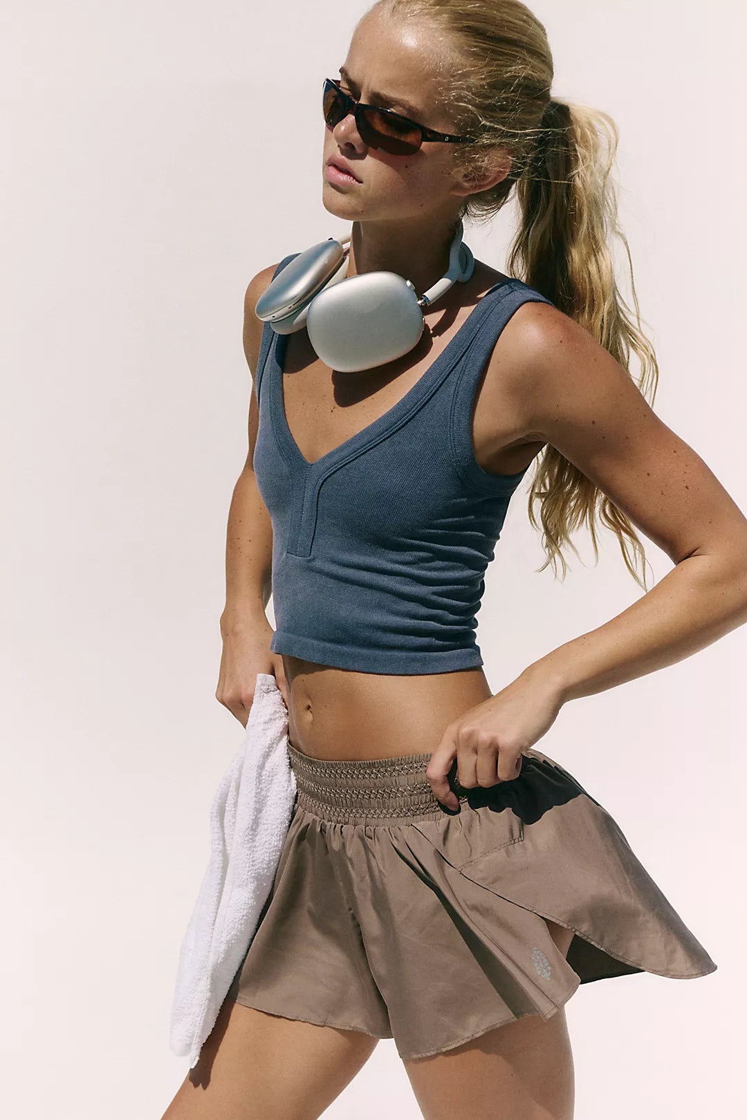 A woman in athletic wear posing against a light beige background, wearing a slate blue cropped tank top, a brown pleated tennis skirt, and silver headphones around her neck.