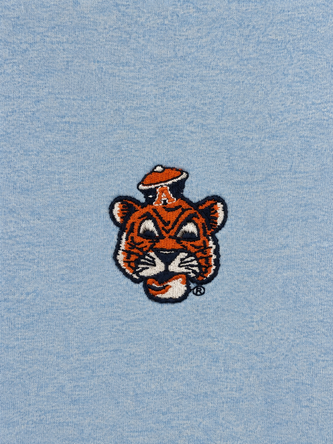 A close-up of a light blue heathered fabric featuring an embroidered tiger mascot head with a small orange hat on its head.