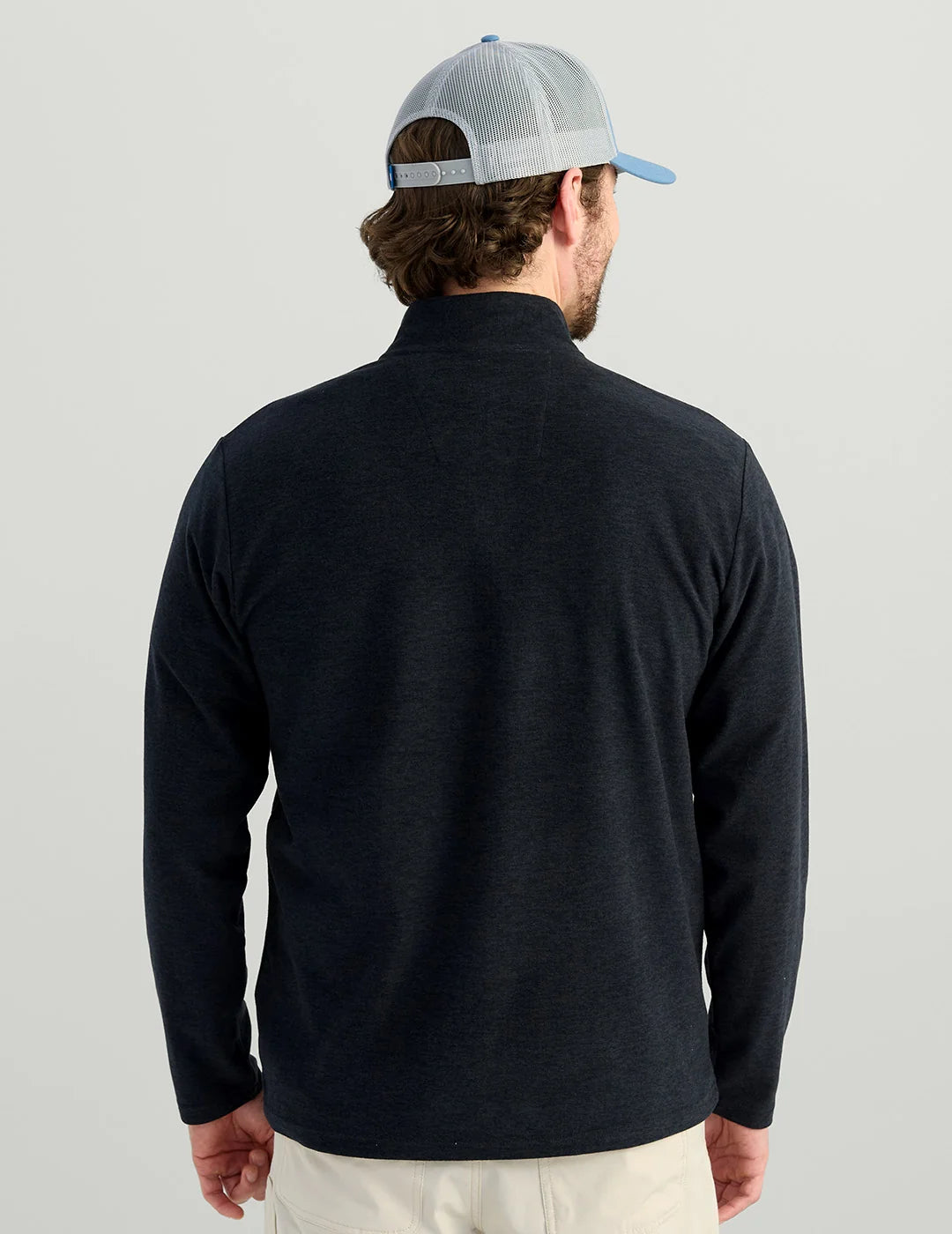 A man viewed from behind wearing a dark navy long-sleeve quarter-zip pullover and a light blue and gray trucker hat, set against a plain light gray studio background.