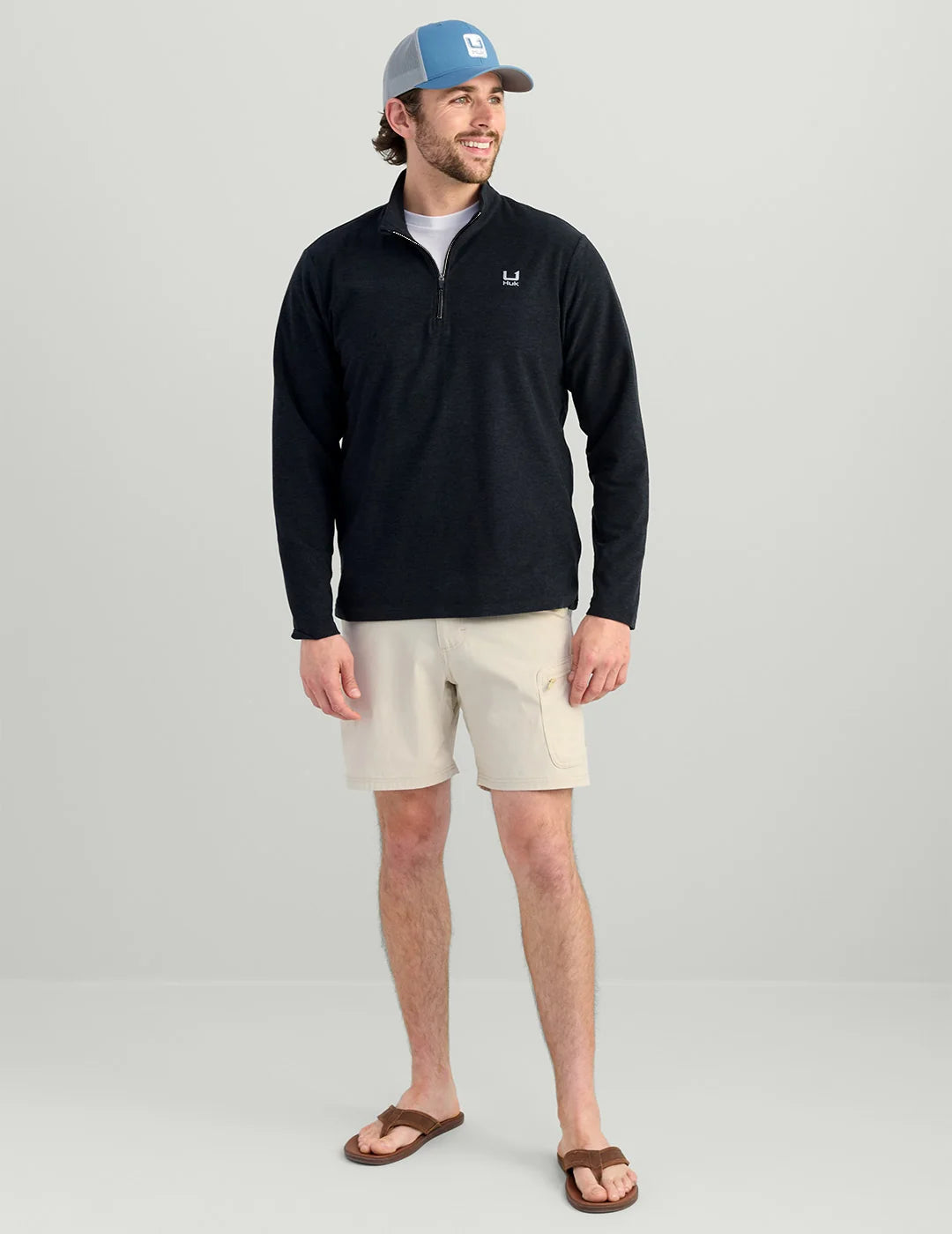 A man modeling a black long-sleeve quarter-zip pullover with a small white chest logo, paired with beige cargo shorts, brown sandals, and a blue trucker hat, against a light gray studio background.