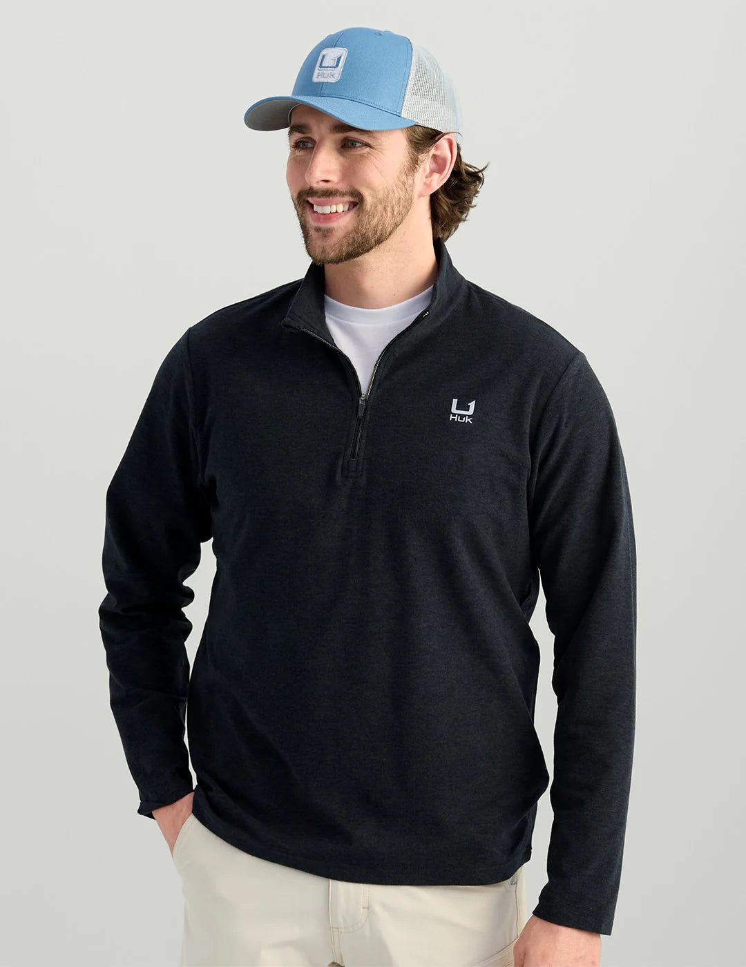 A man wearing a dark navy quarter-zip long-sleeve pullover with a small white logo on the left chest, paired with a light blue and gray trucker hat and off-white pants, against a solid light gray background.