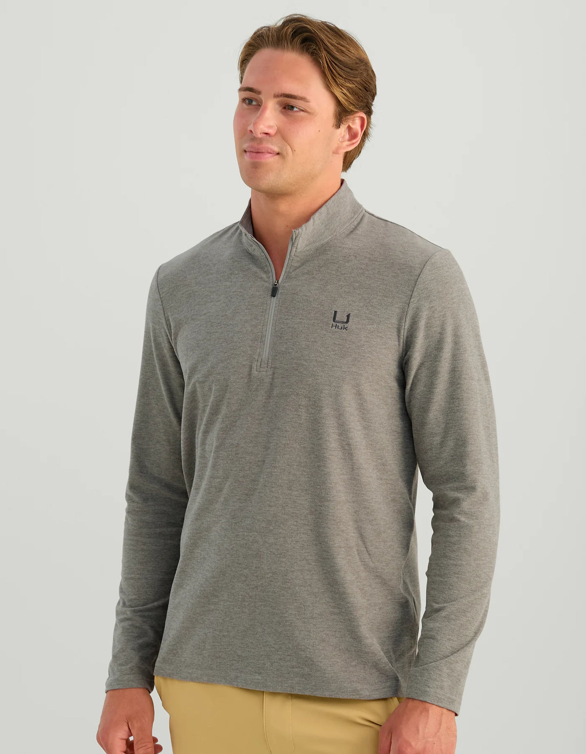 A man wearing a heather gray long-sleeve quarter-zip pullover with a small dark embroidered logo on the left chest, paired with tan pants, against a solid light gray background.