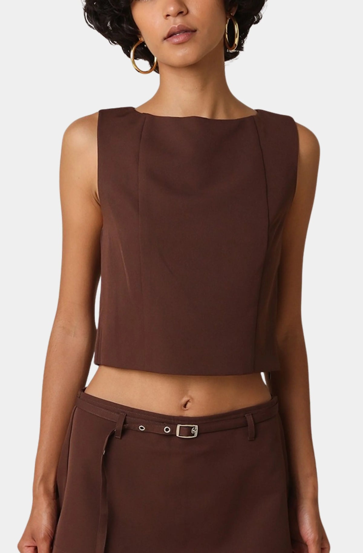 A model wearing a sleeveless, boat-neck cropped top in a dark brown color, paired with matching brown bottoms and gold hoop earrings, against a light gray background.