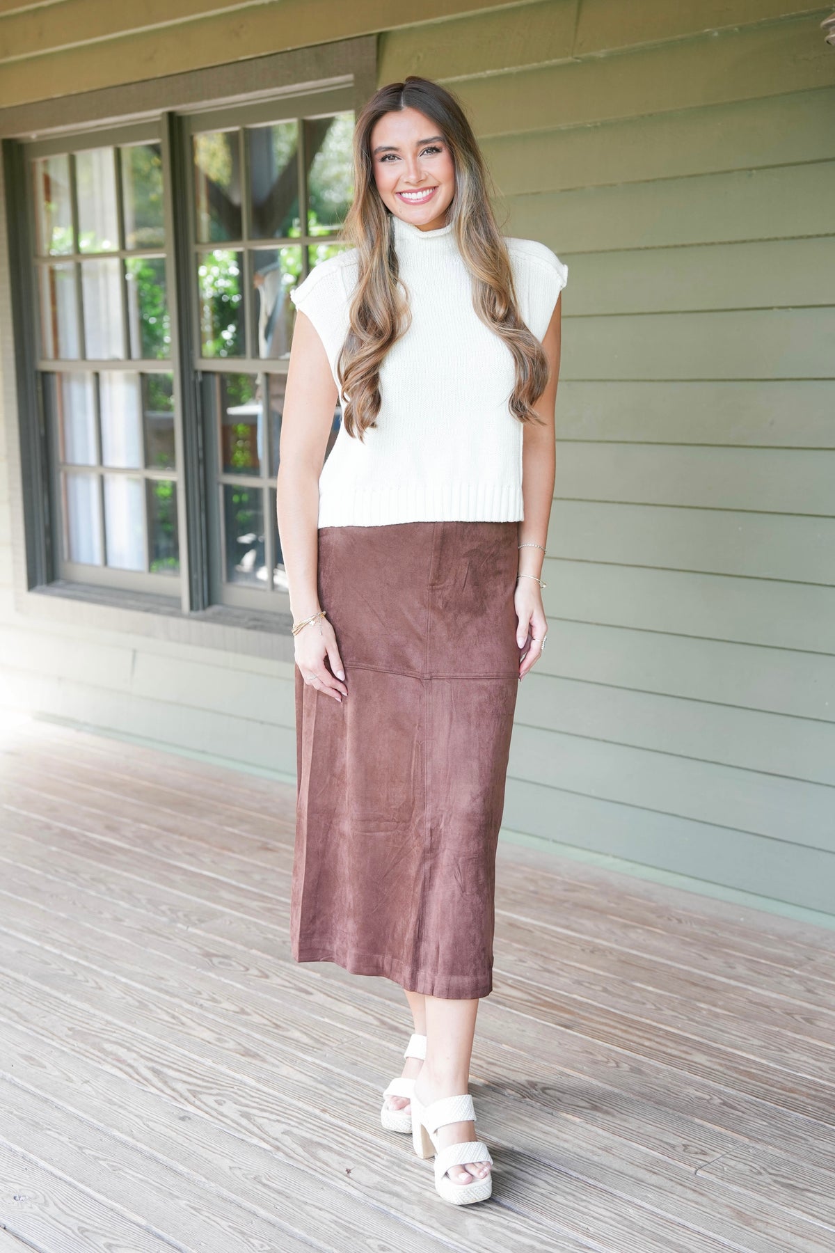 A woman stands on a wooden porch wearing a white ribbed knit mock-neck top and a brown midi-length suede skirt, paired with white platform sandals.