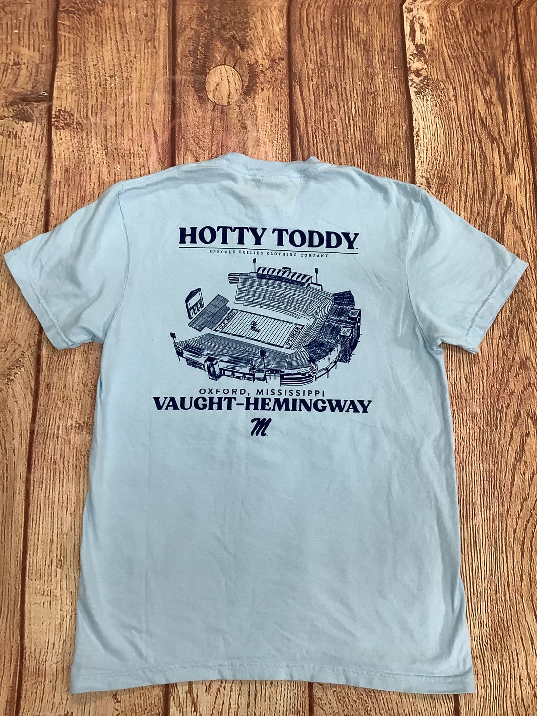 A light blue short-sleeve t-shirt laid flat on a rustic wooden plank background, showing the back side with a dark blue graphic of a stadium and text reading 'HOTTY TODDY' and 'VAUGHT-HEMINGWAY'.