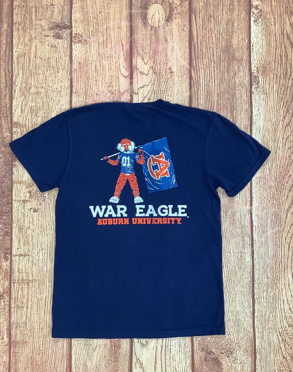 A navy blue short-sleeve t-shirt featuring a graphic of the Auburn University War Eagle mascot holding a blue flag with an orange 'AU' logo, with 'WAR EAGLE' and 'AUBURN UNIVERSITY' printed in white and orange below. The shirt is laid flat on a rustic wooden plank background.