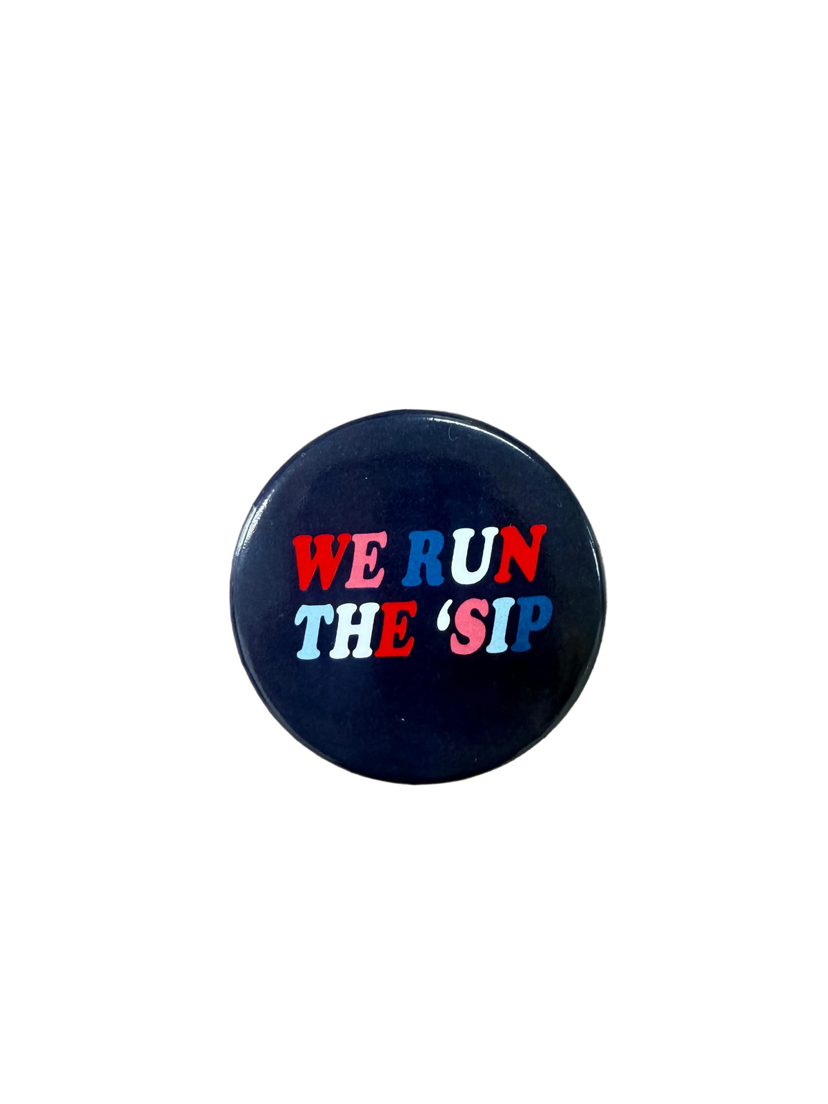 A round black button badge with the text 'WE RUN THE 'SIP' in red, white, and blue lettering, placed on a light brown wooden surface.