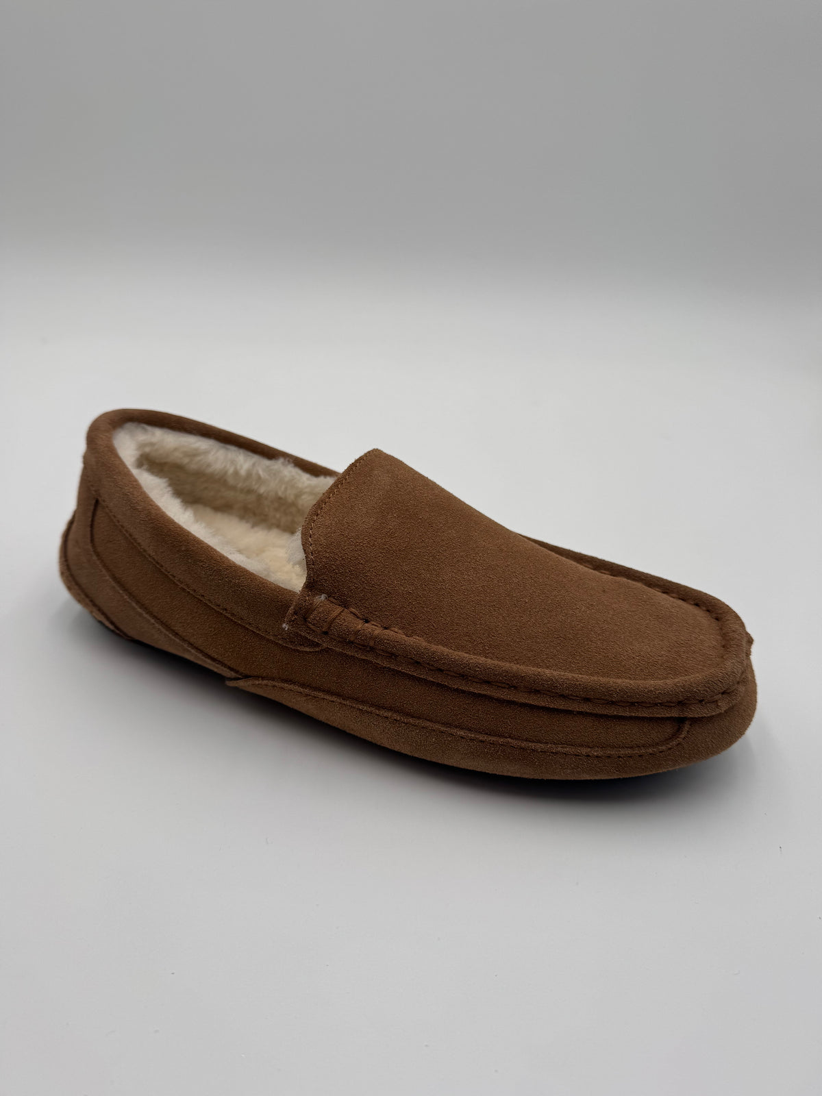 A single brown suede slip-on slipper with a soft cream-colored shearling lining, photographed on a neutral gray studio background.