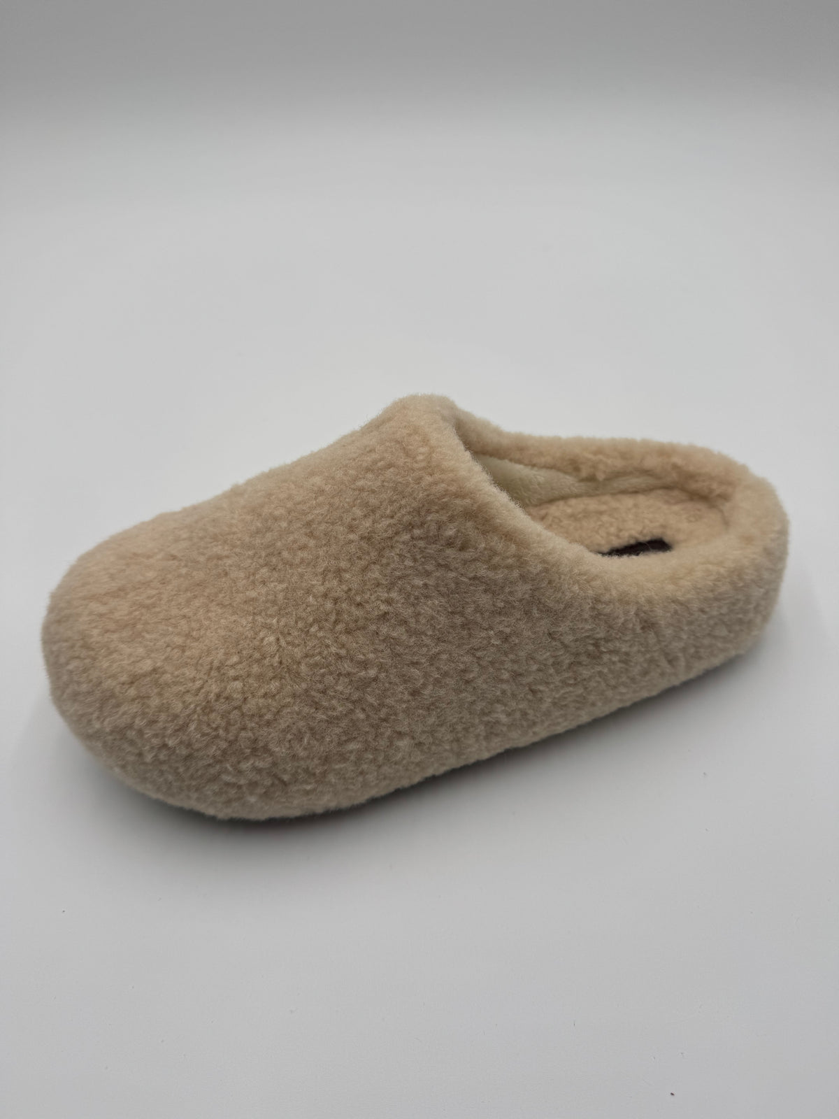 A single fuzzy slip-on mule slipper in a light tan or beige color, photographed on a clean light gray studio background.