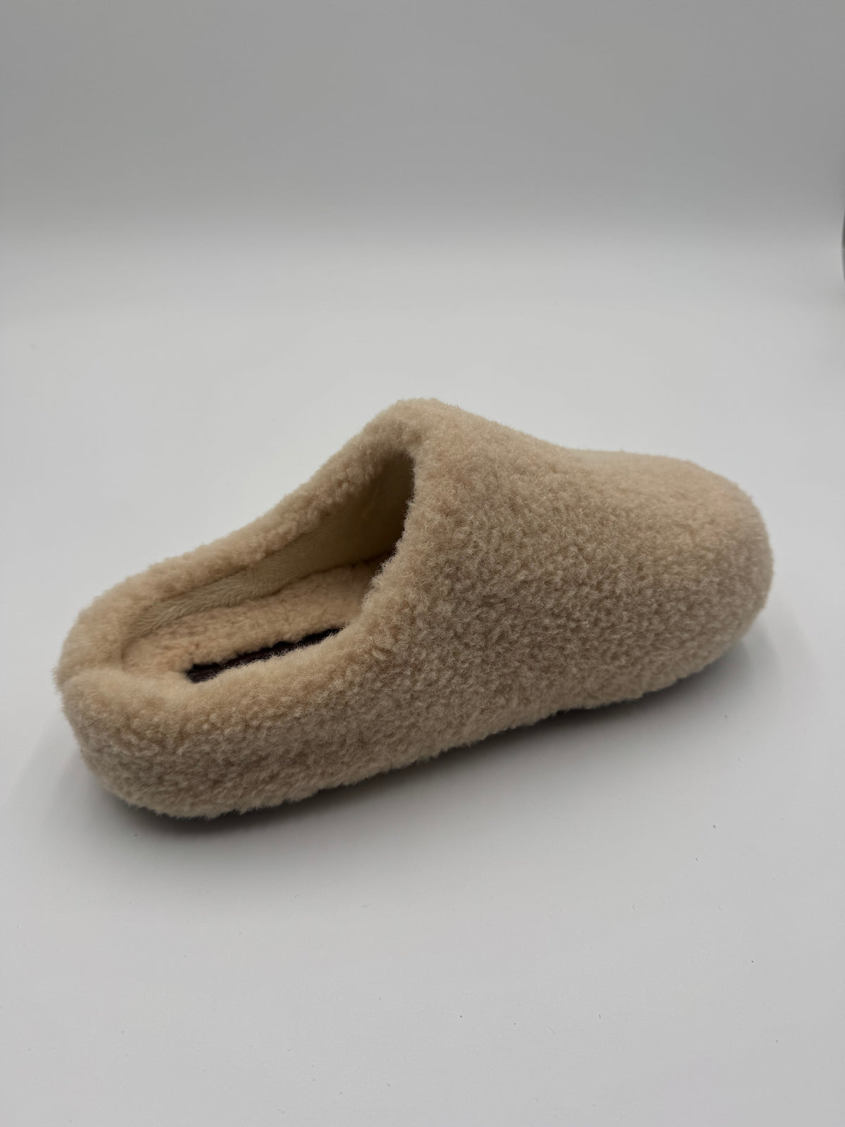 A single beige fuzzy slipper or clog-style house shoe photographed on a plain light gray studio background.