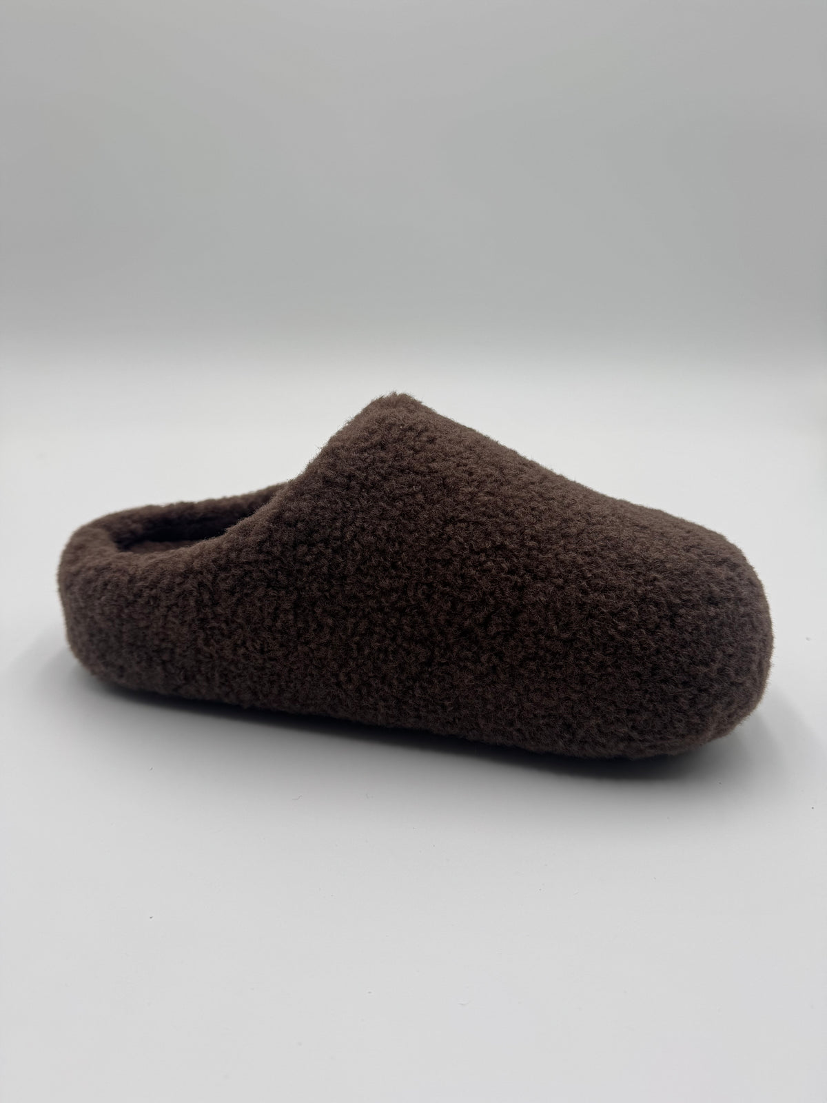 A single dark brown fuzzy slip-on slipper photographed on a plain light gray studio background.