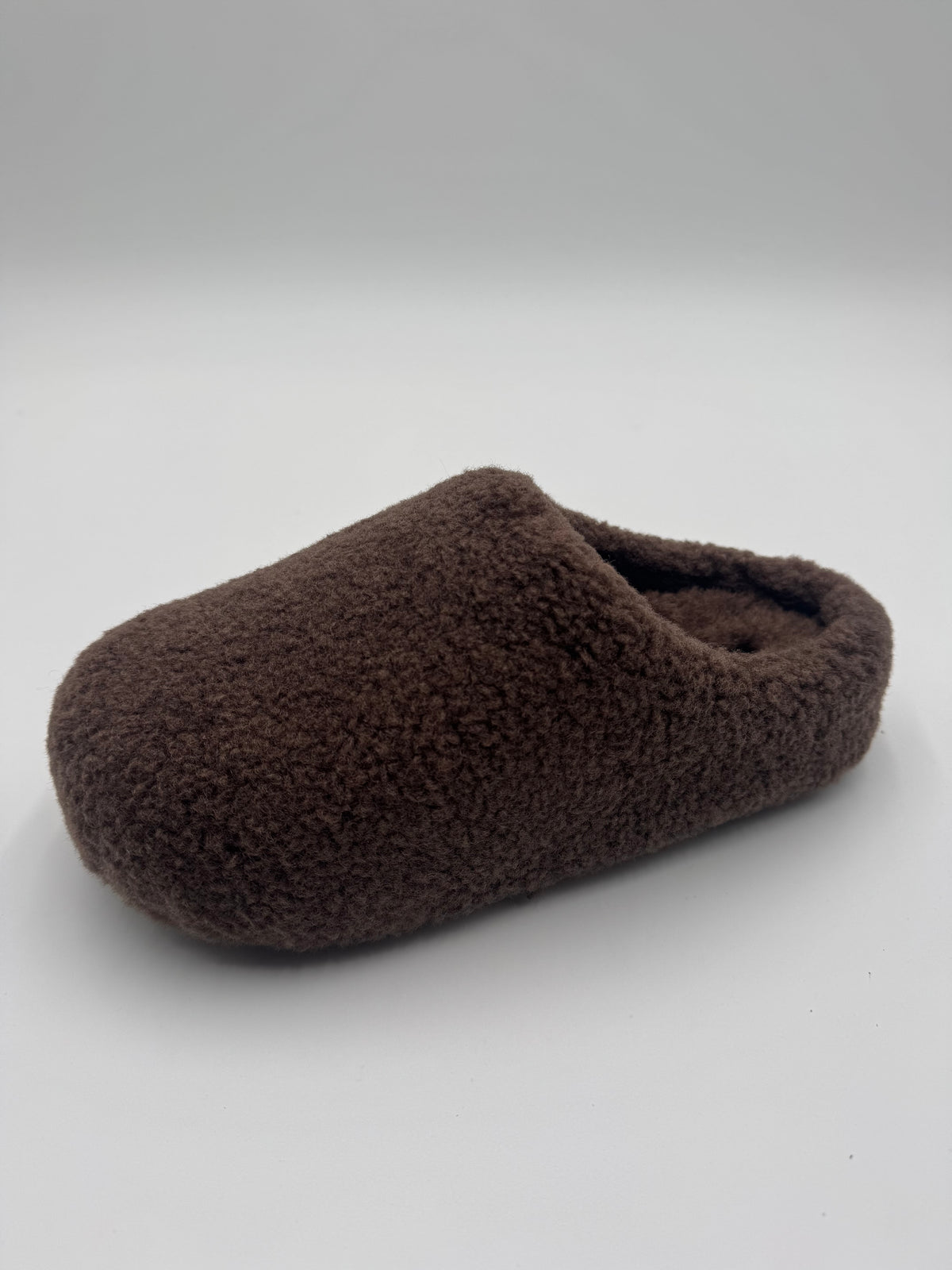 A single dark brown fuzzy slipper or clog-style house shoe photographed on a plain light gray studio background.