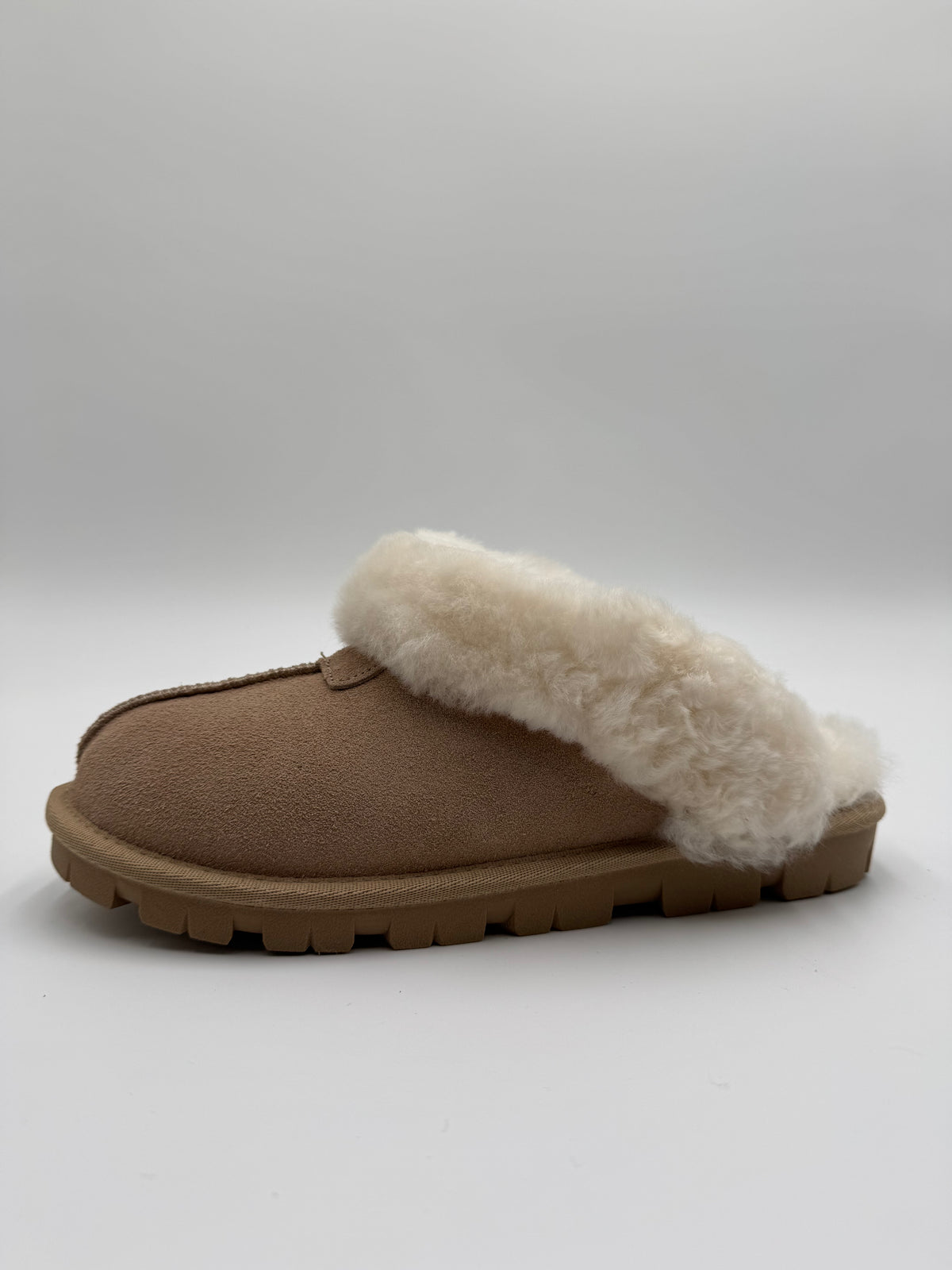 A single slip-on mule slipper in tan suede with a thick, fluffy cream shearling lining and a brown lug sole, shown against a neutral gray background.