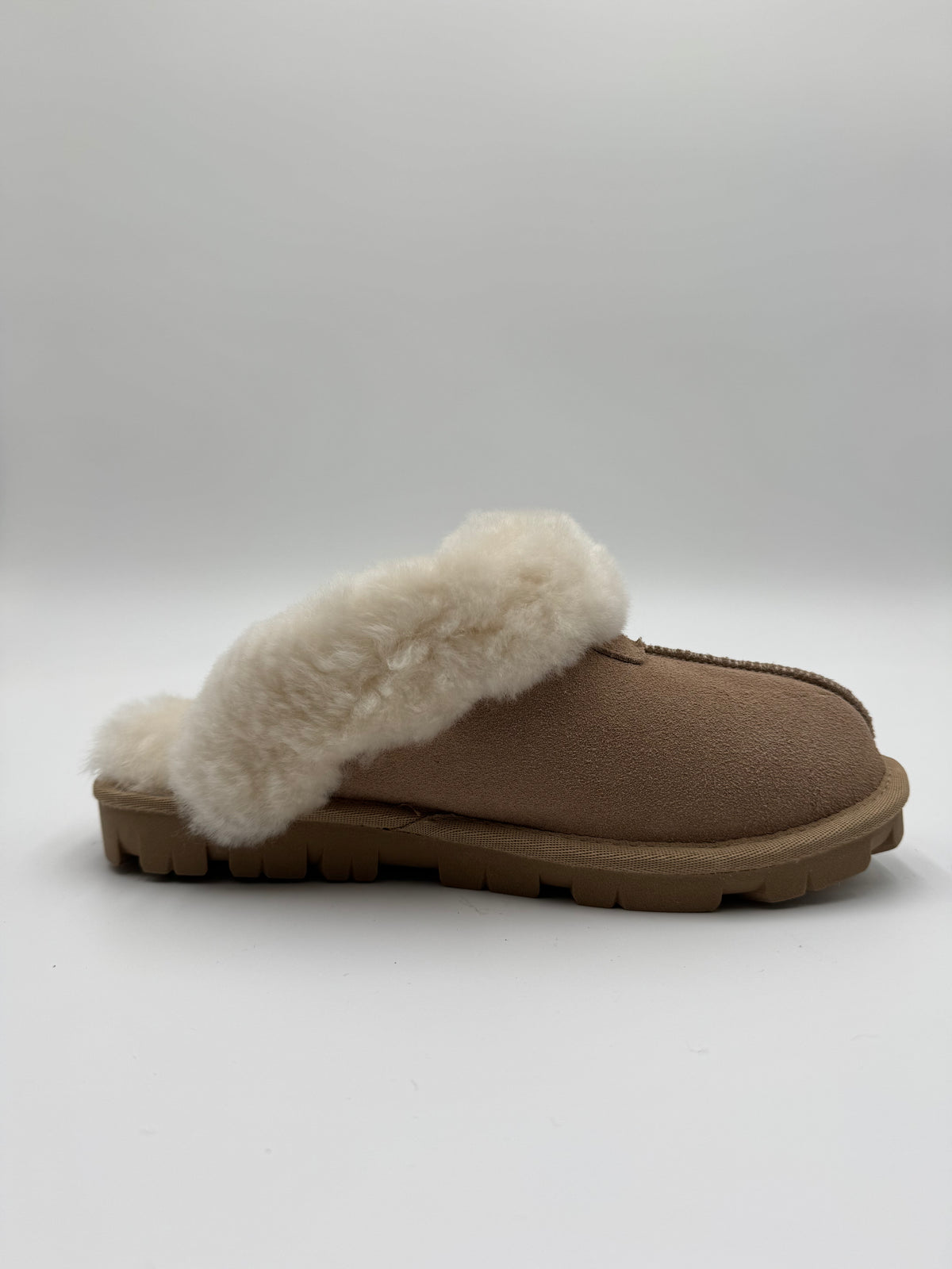 A single slip-on mule slipper in tan faux suede with a thick, fluffy cream shearling-style collar and a brown sole, shown from a side profile on a light gray background.