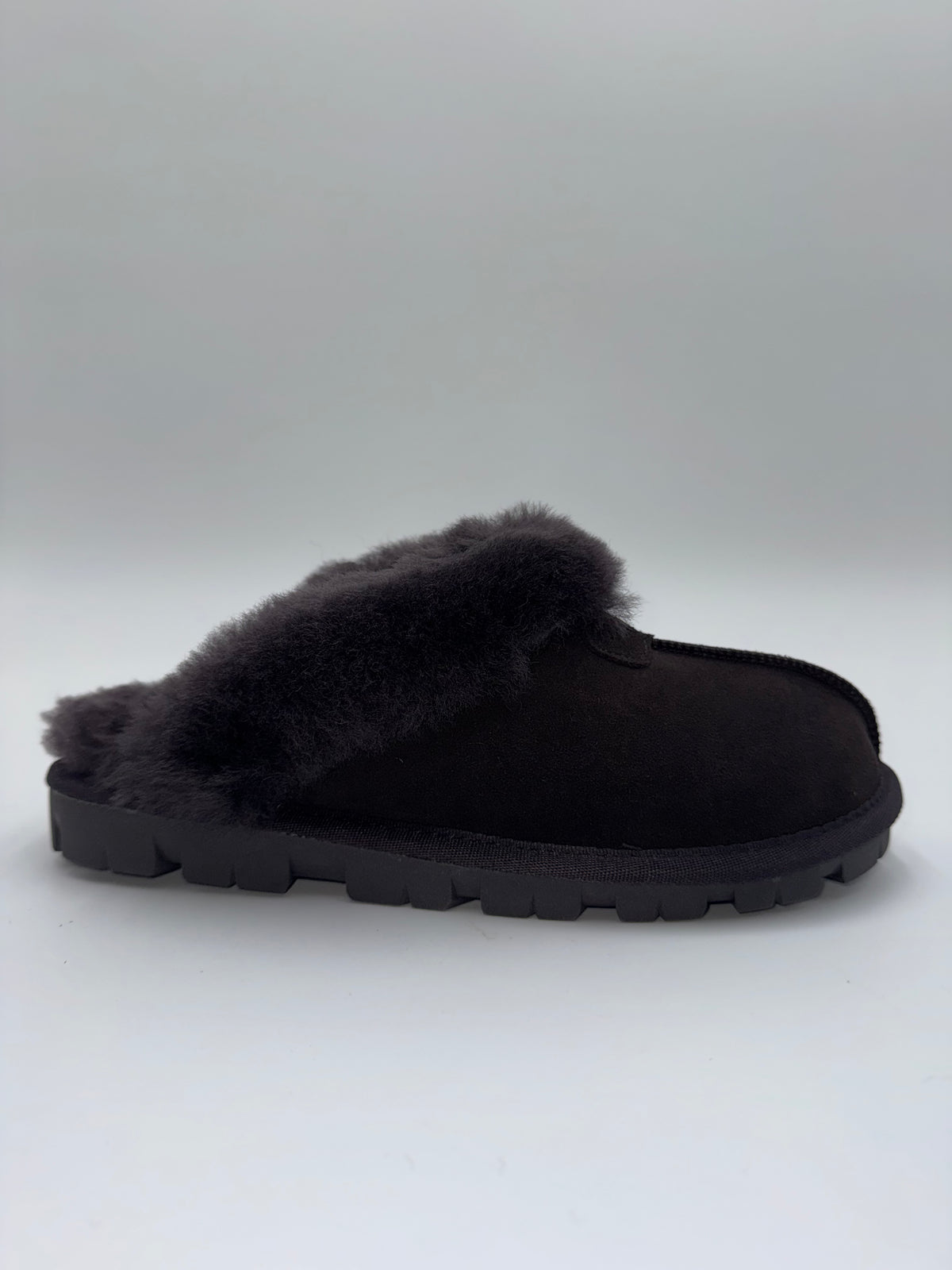 A single dark brown slip-on mule slipper with a thick black sole and dark gray fluffy shearling lining, shown from a side profile against a light gray background.