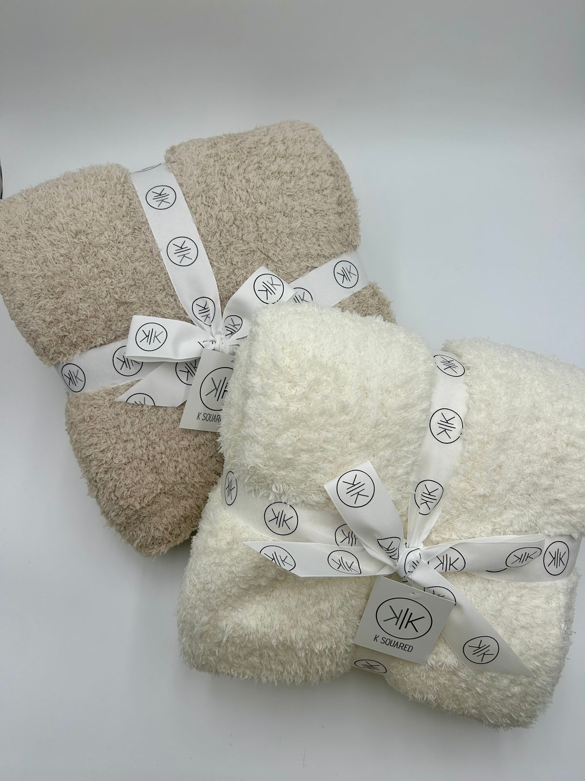 Two plush fleece throw blankets, one in beige and one in cream, wrapped in white ribbons with a black monogram logo, on a light gray background.
