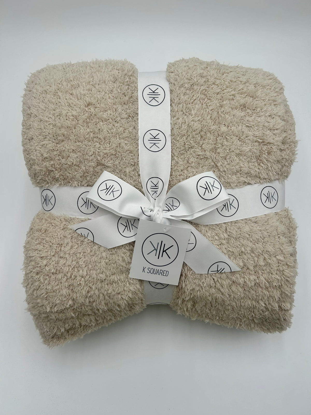A folded beige terry cloth towel or blanket wrapped with a white ribbon printed with the K SQUARED logo, set against a plain light gray background.